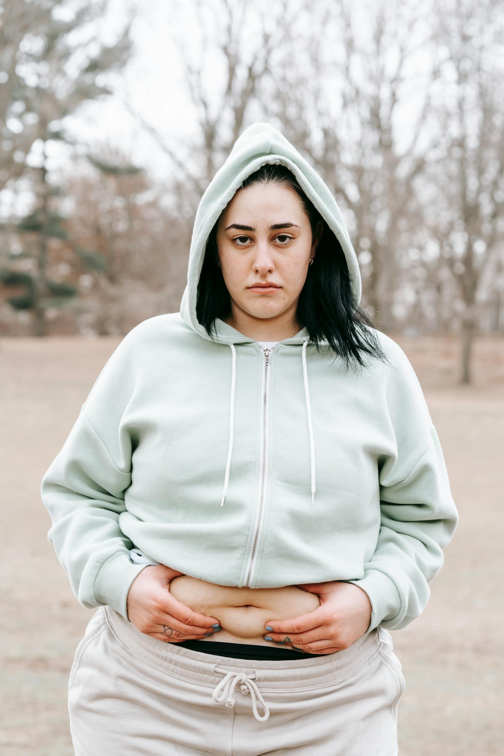 Gloomy female in sportswear showing folds on waist and looking at camera while doing exercises in autumn day