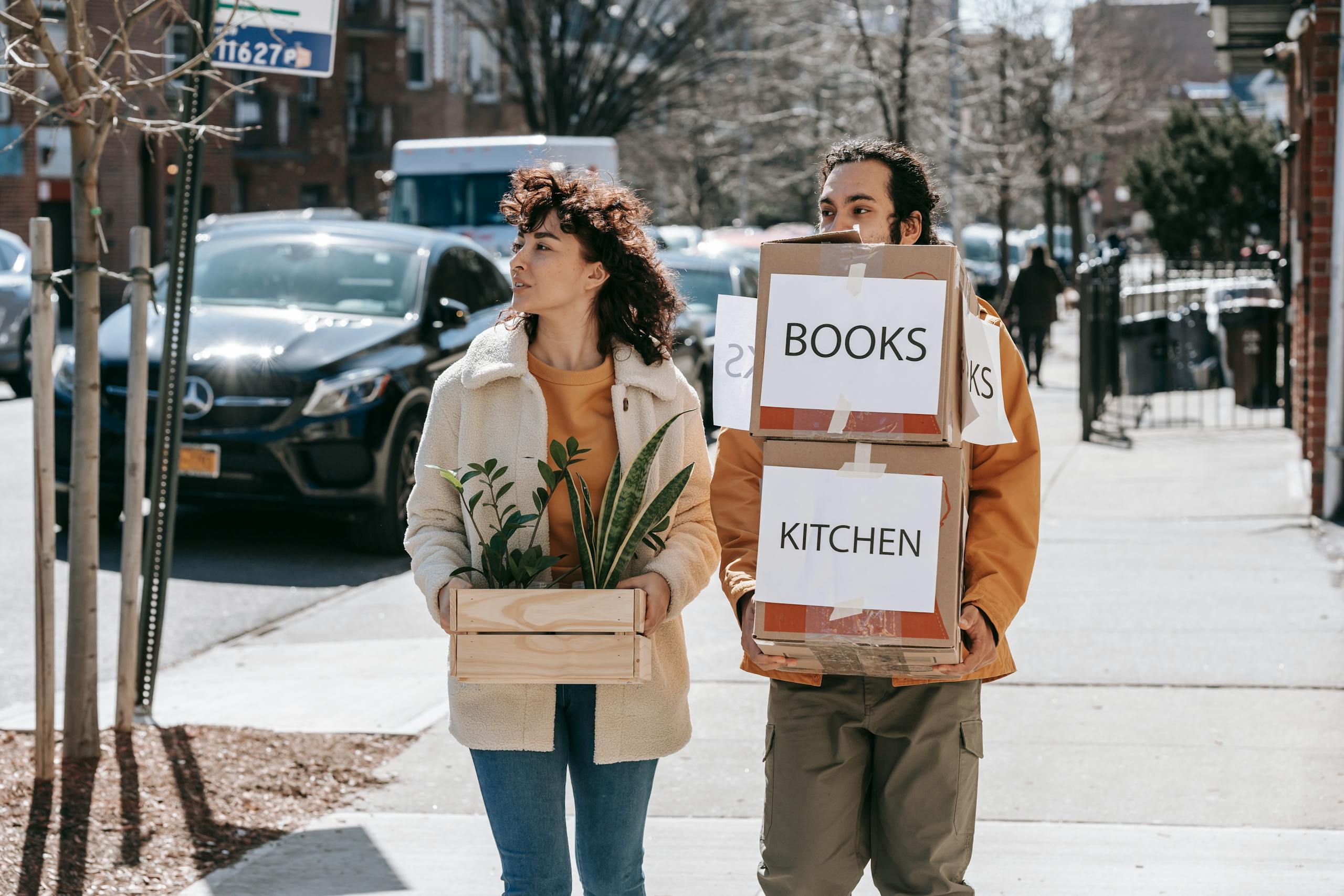 A couple walks in a city street carrying boxes labeled 'Books' and 'Kitchen' symbolizing relocation.