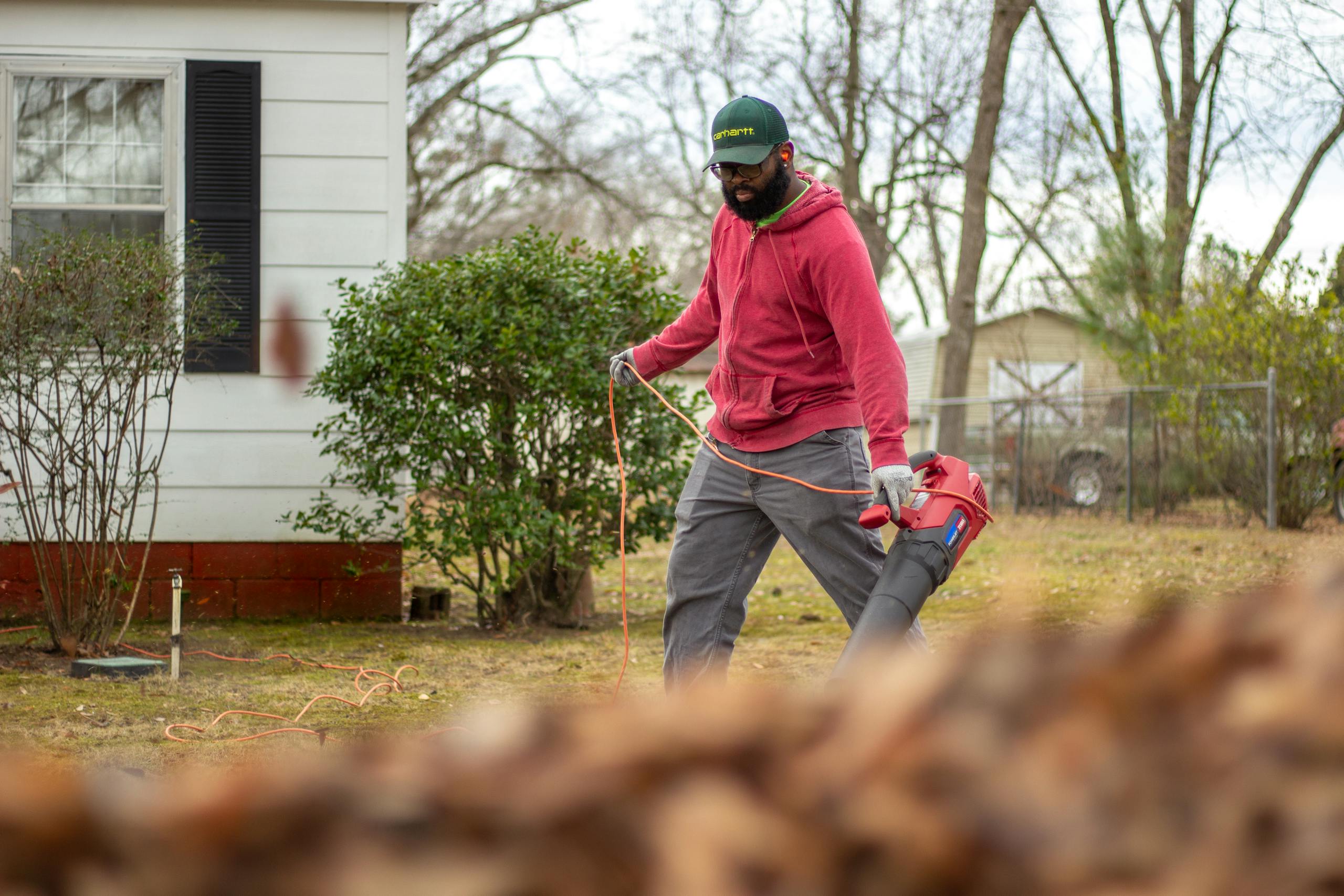 A man in a red jacket operates a leaf blower in an autumn backyard, clearing leaves.