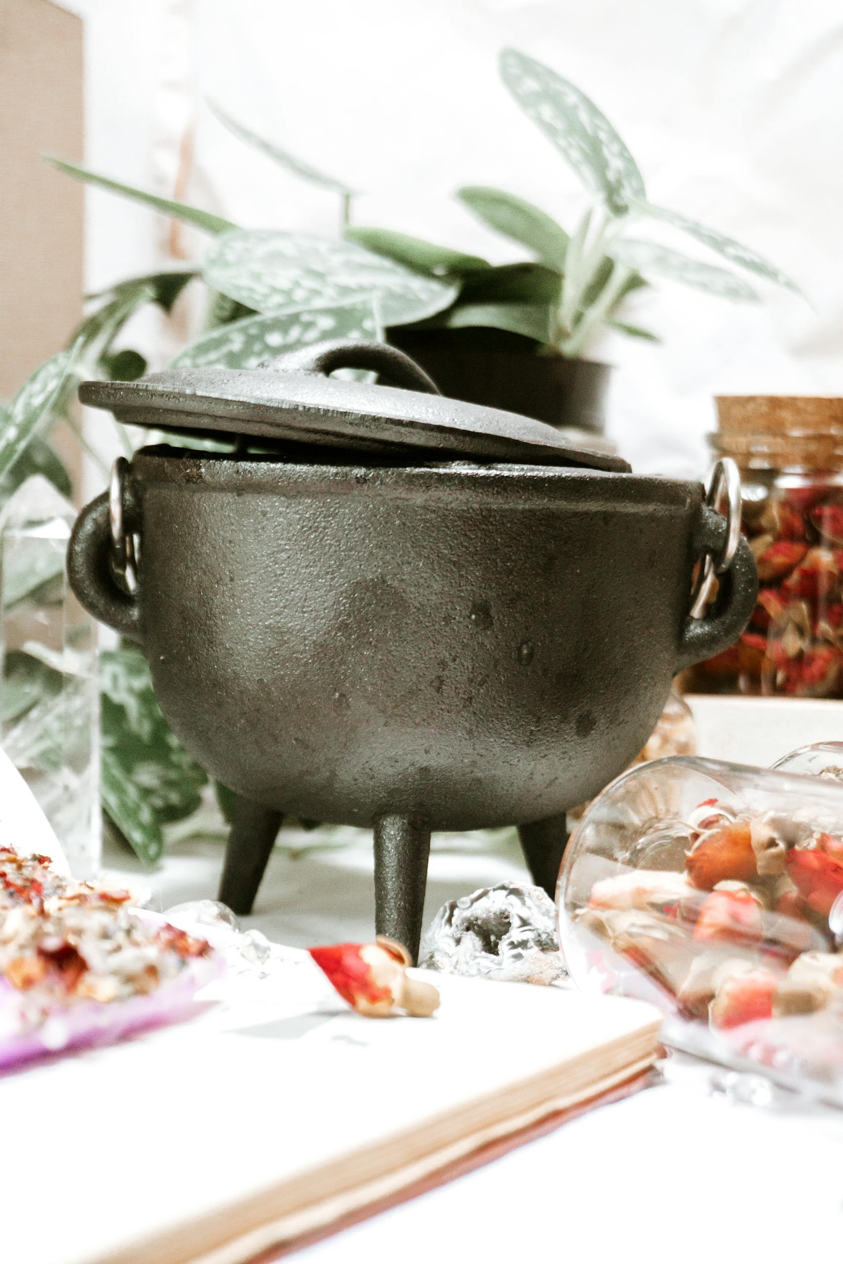 A rustic cast iron cauldron surrounded by plants and ingredients in a cozy kitchen.
