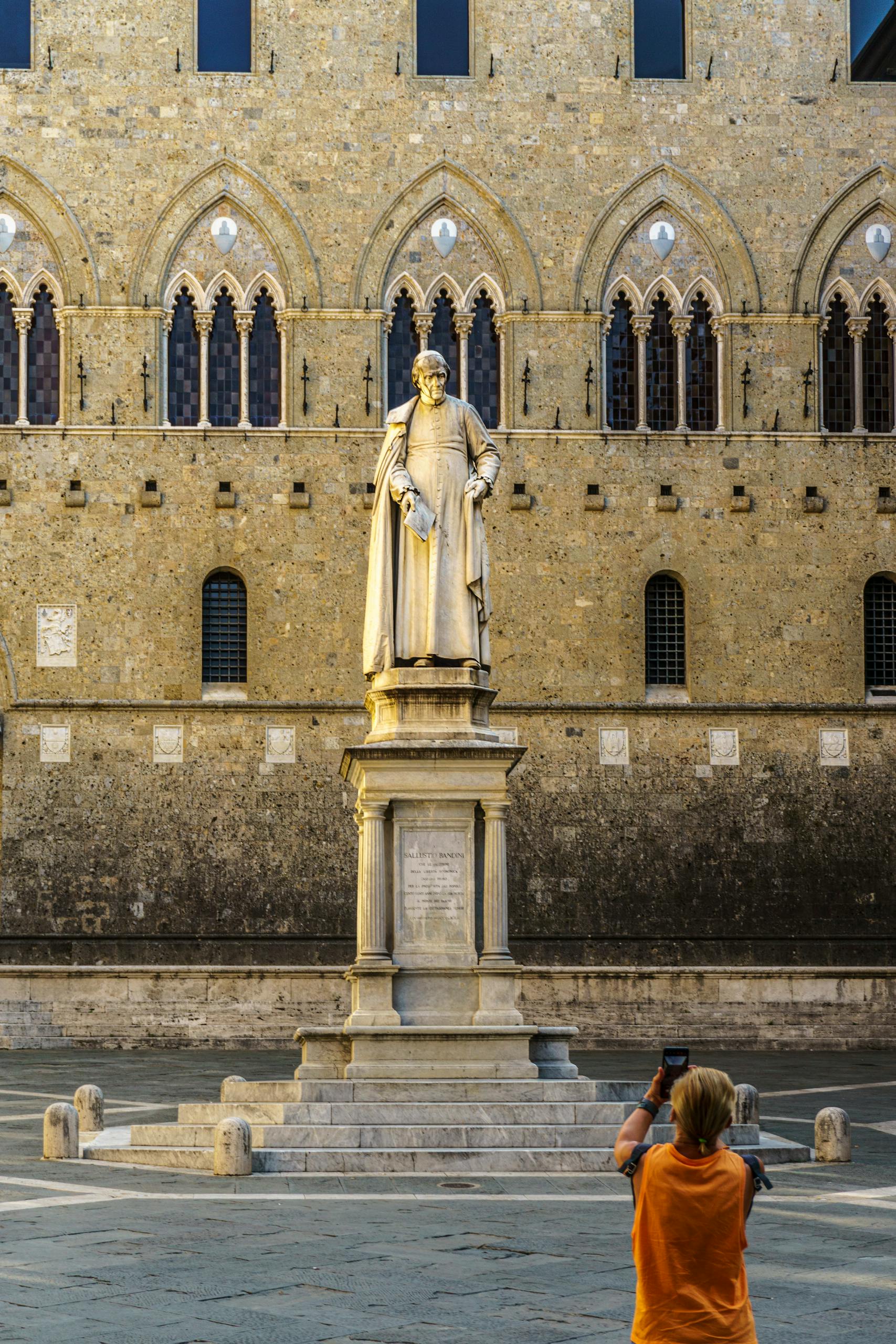 A tourist captures a moment at the Piazza Salimbeni with a historic statue in Siena, Italy.