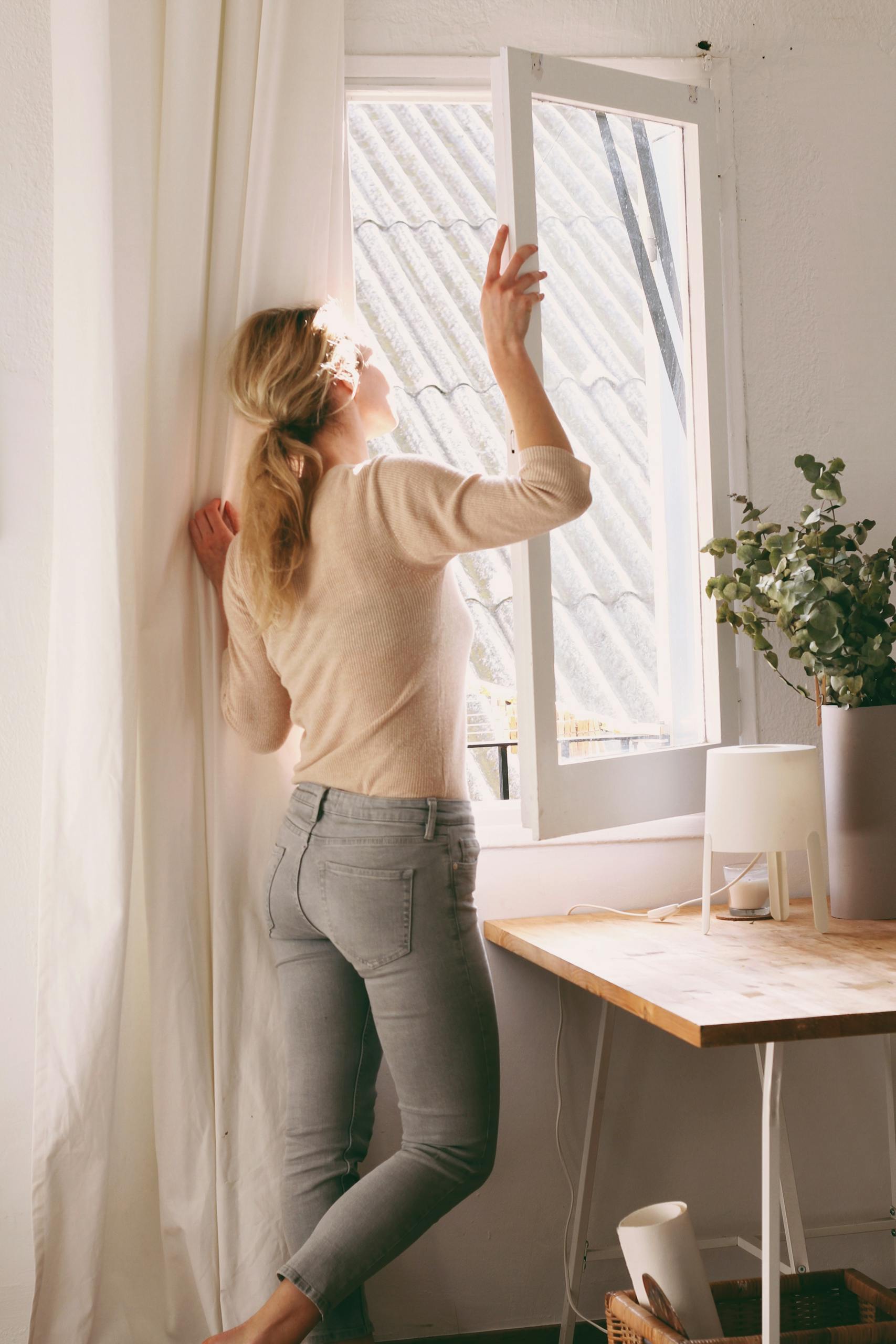 A woman savoring the sunlight through an open window in a cozy, minimalist room.
