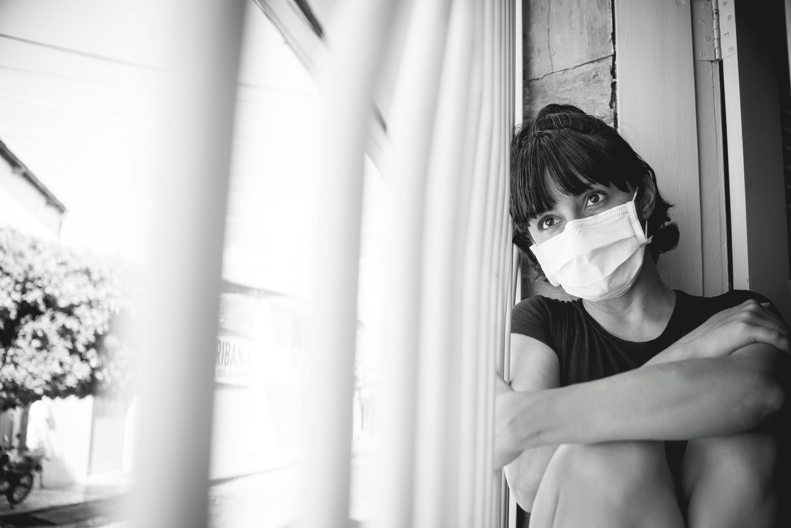 A woman wearing a face mask sits by a window during quarantine, reflecting solitude.