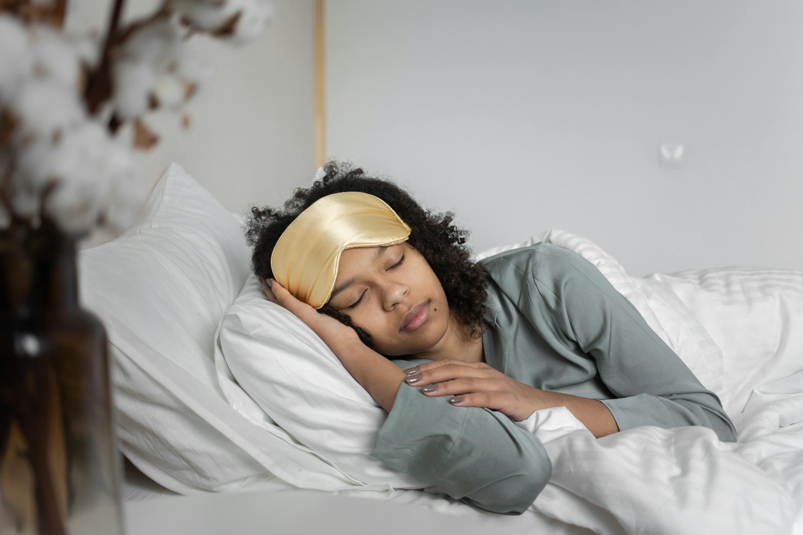 A young woman peacefully sleeps with a sleep mask, nestled in cozy white bedding.