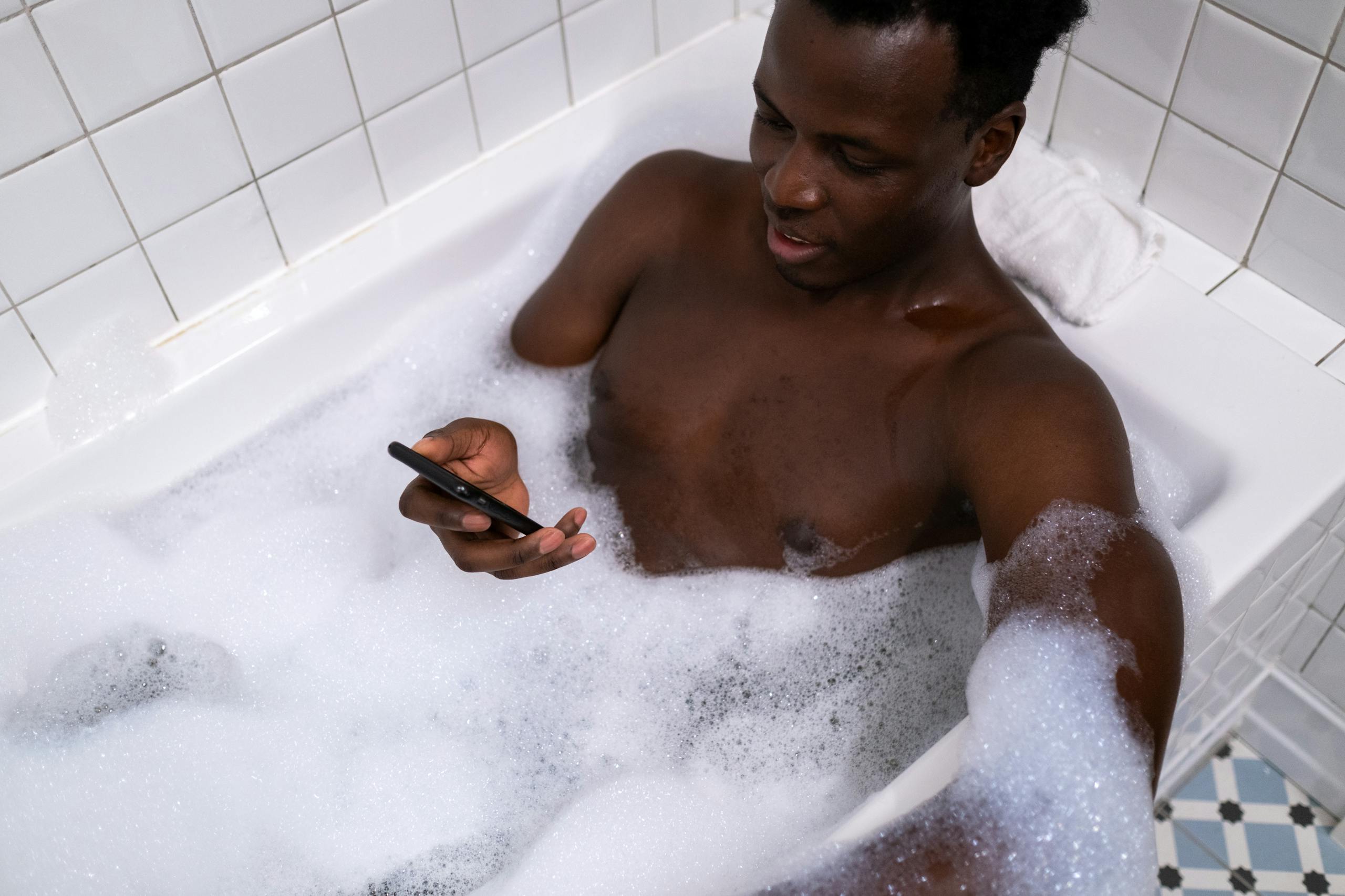 An adult man enjoying a bubble bath while using a mobile phone in a white tiled bathroom.