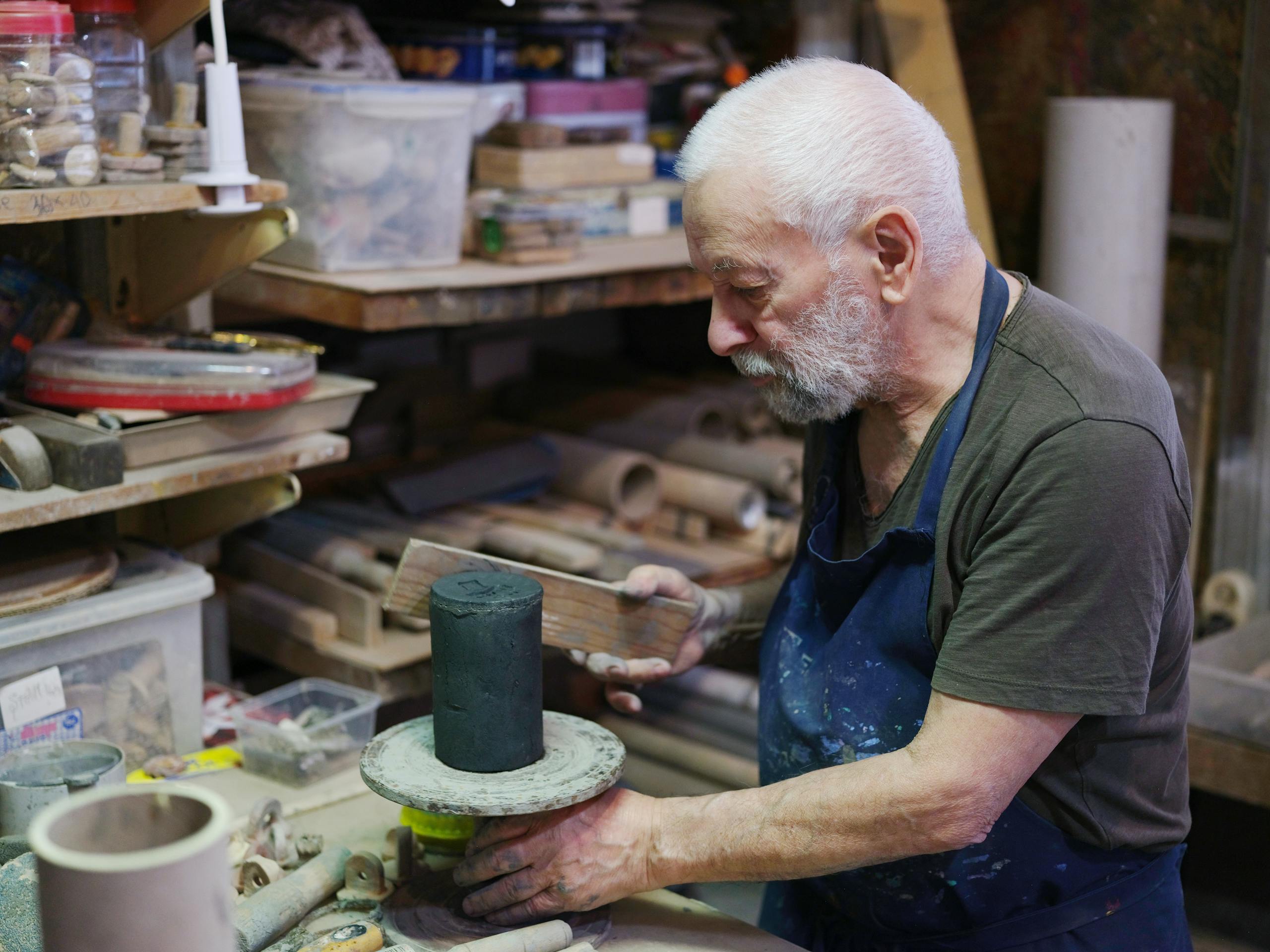 An elderly man focuses on crafting in a cluttered workshop, highlighting skilled craftsmanship.