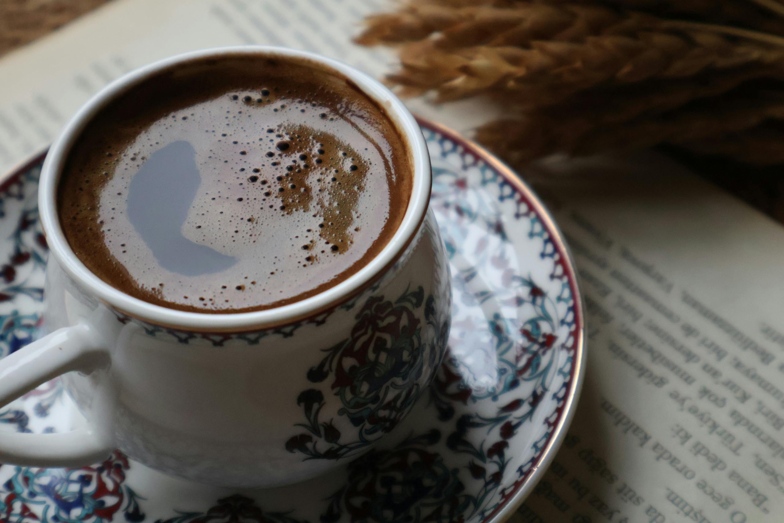 Close-up of Turkish coffee in an ornate cup next to wheat and an open book.
