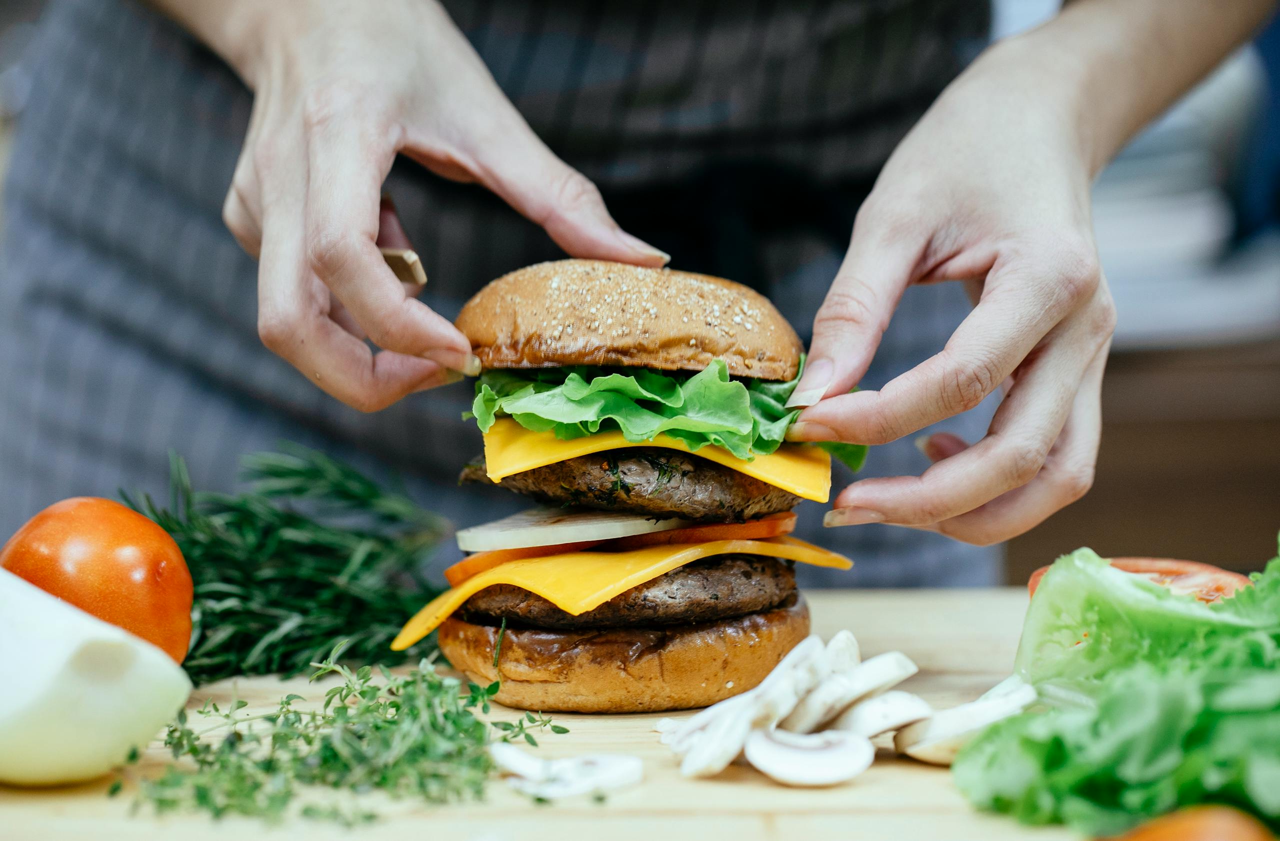 Crop anonymous female cook putting soft bread bun on top of lettuce leaves and meat patty while preparing double cheeseburger in kitchen