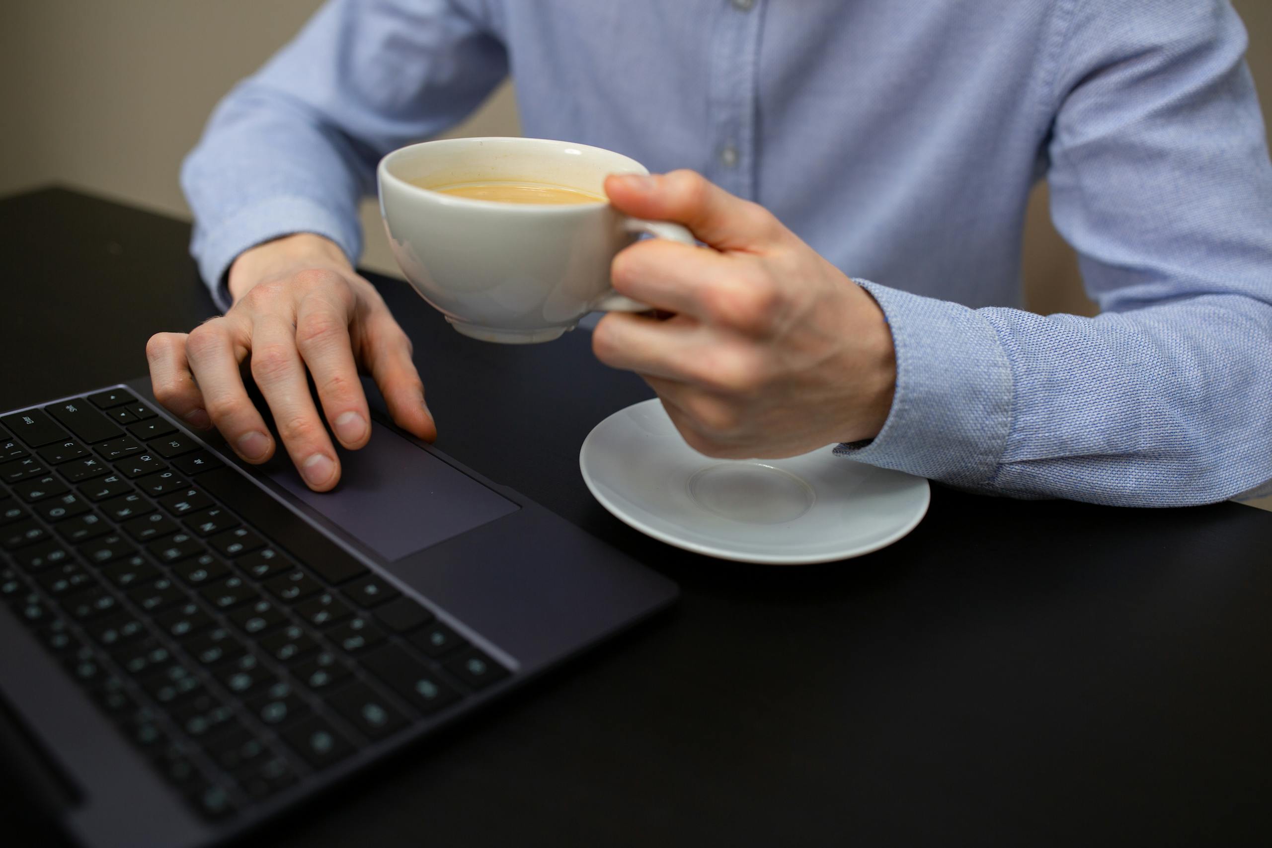 Crop unrecognizable male entrepreneur drinking hot cappuccino while working on laptop in office