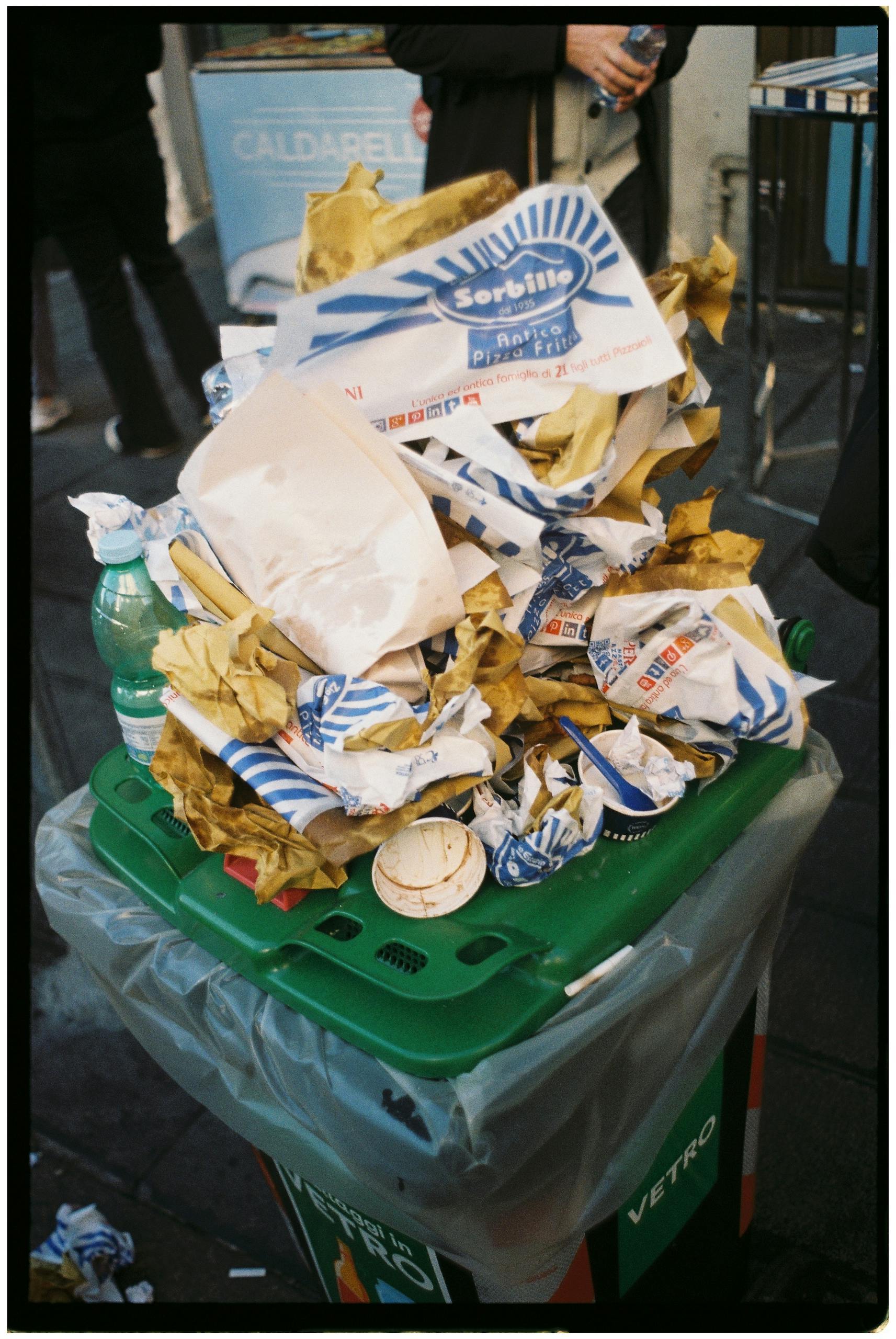 Overflowing trash bin filled with pizza wrappers on a street in Naples, Italy.