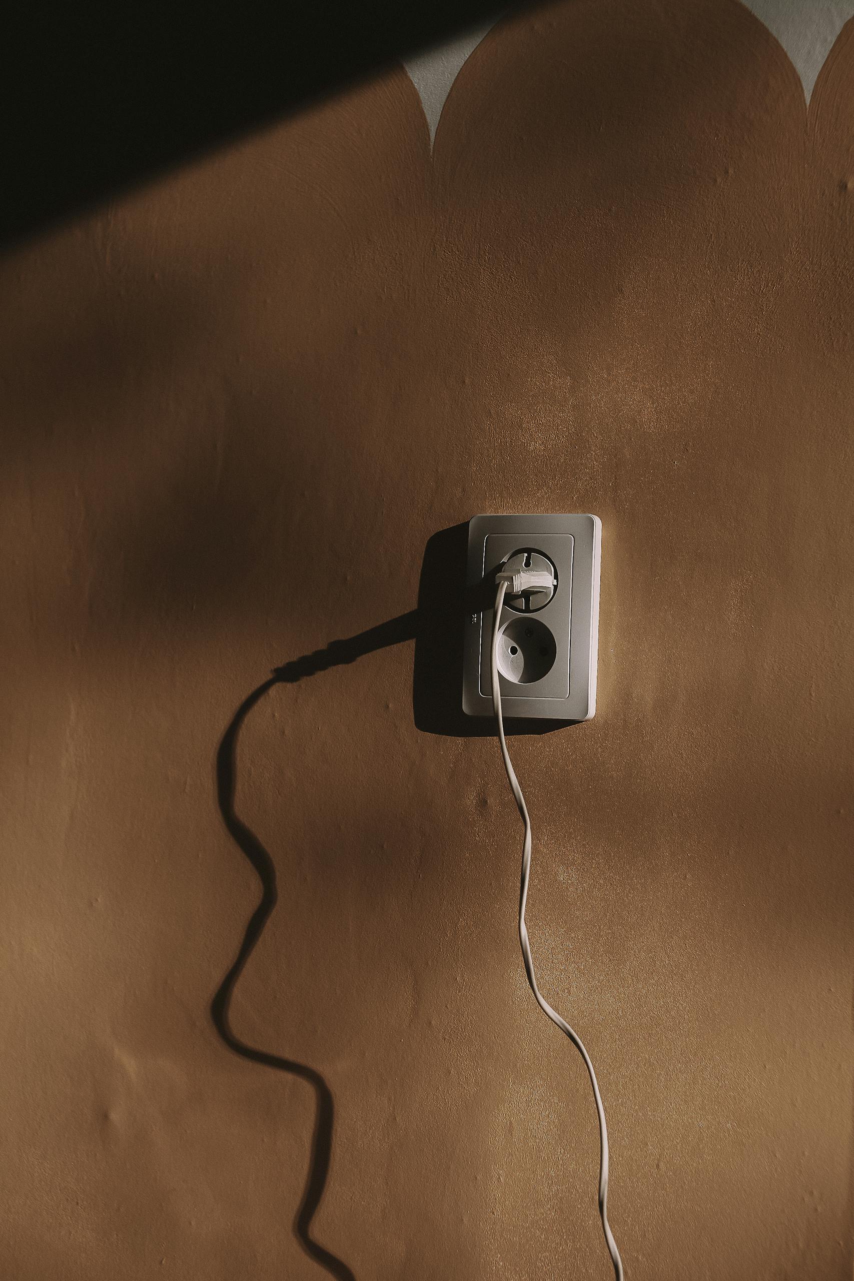 Vertical close-up of a socket with a plugged wire casting dramatic shadows on a textured wall.