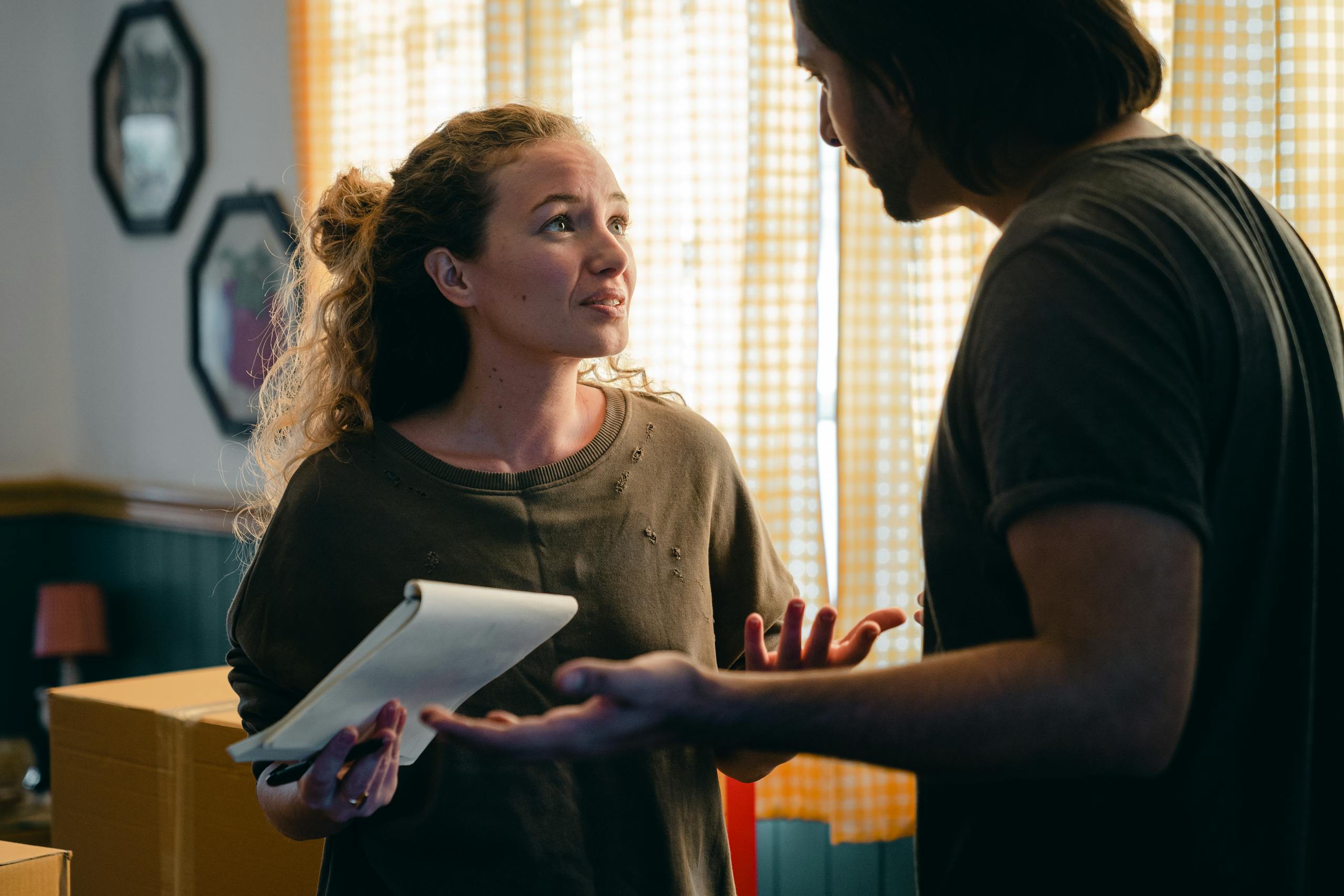 Young perplexed couple wearing casual clothes with notebook looking at each other nervously while unpacking carton box