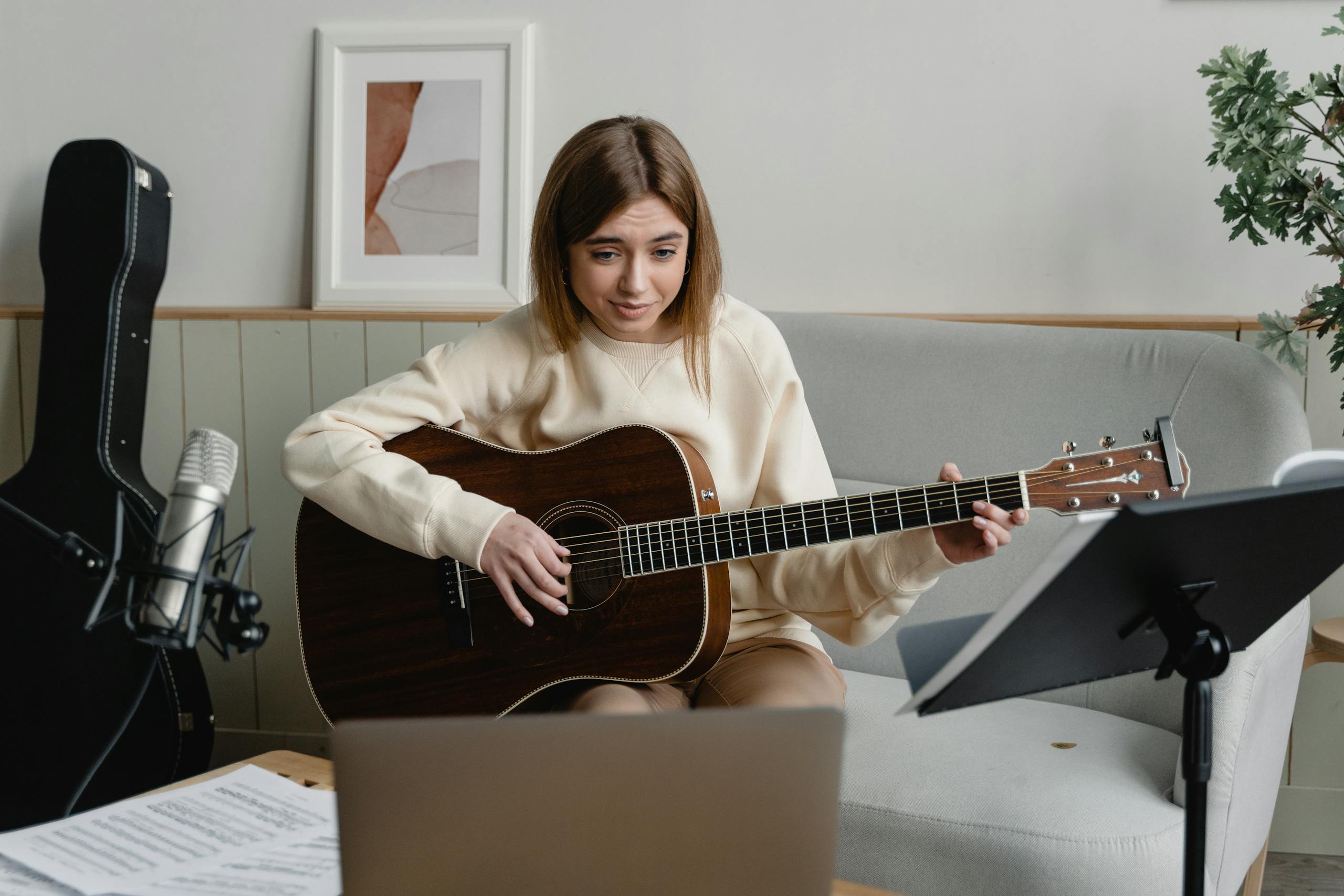 Young woman playing guitar on a sofa while attending an online music lesson at home.