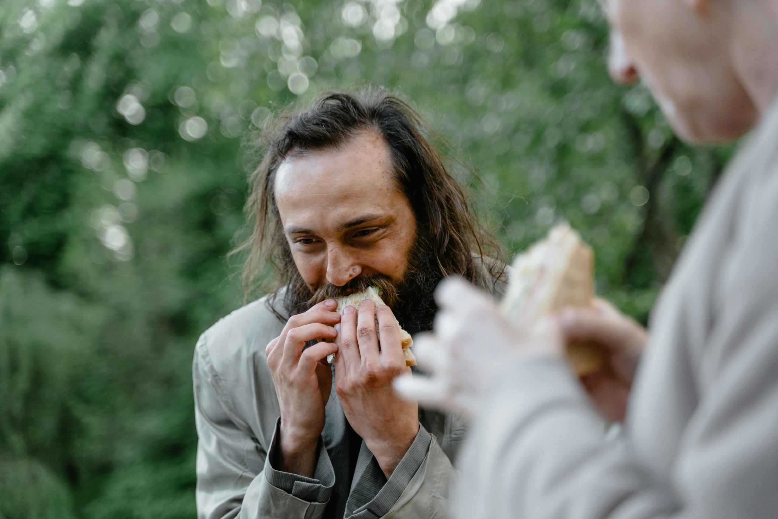 A man happily eats a sandwich outside in a green and natural setting.