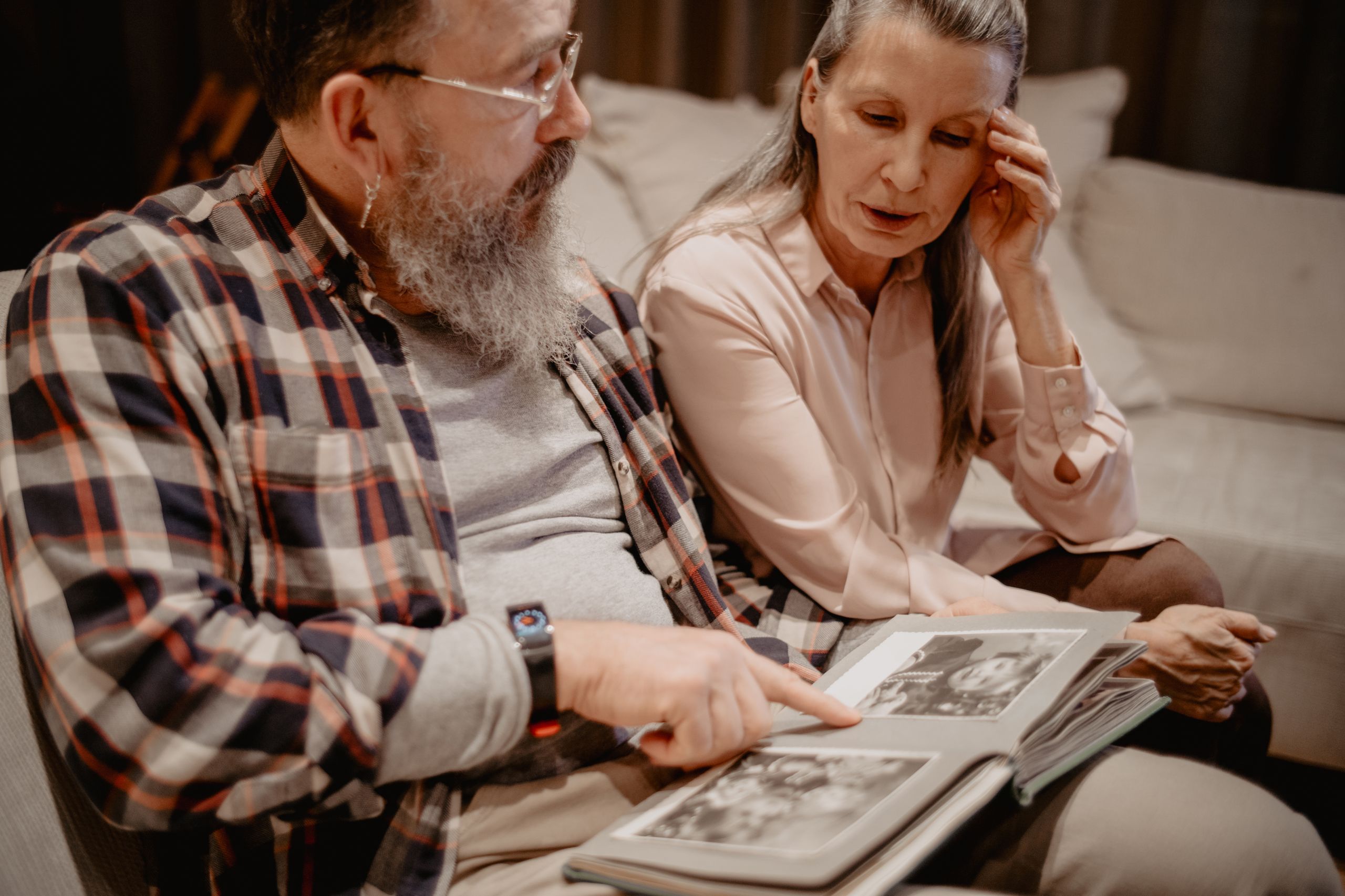 Senior couple sits indoors reminiscing over a photo album, sharing memories and bonding.