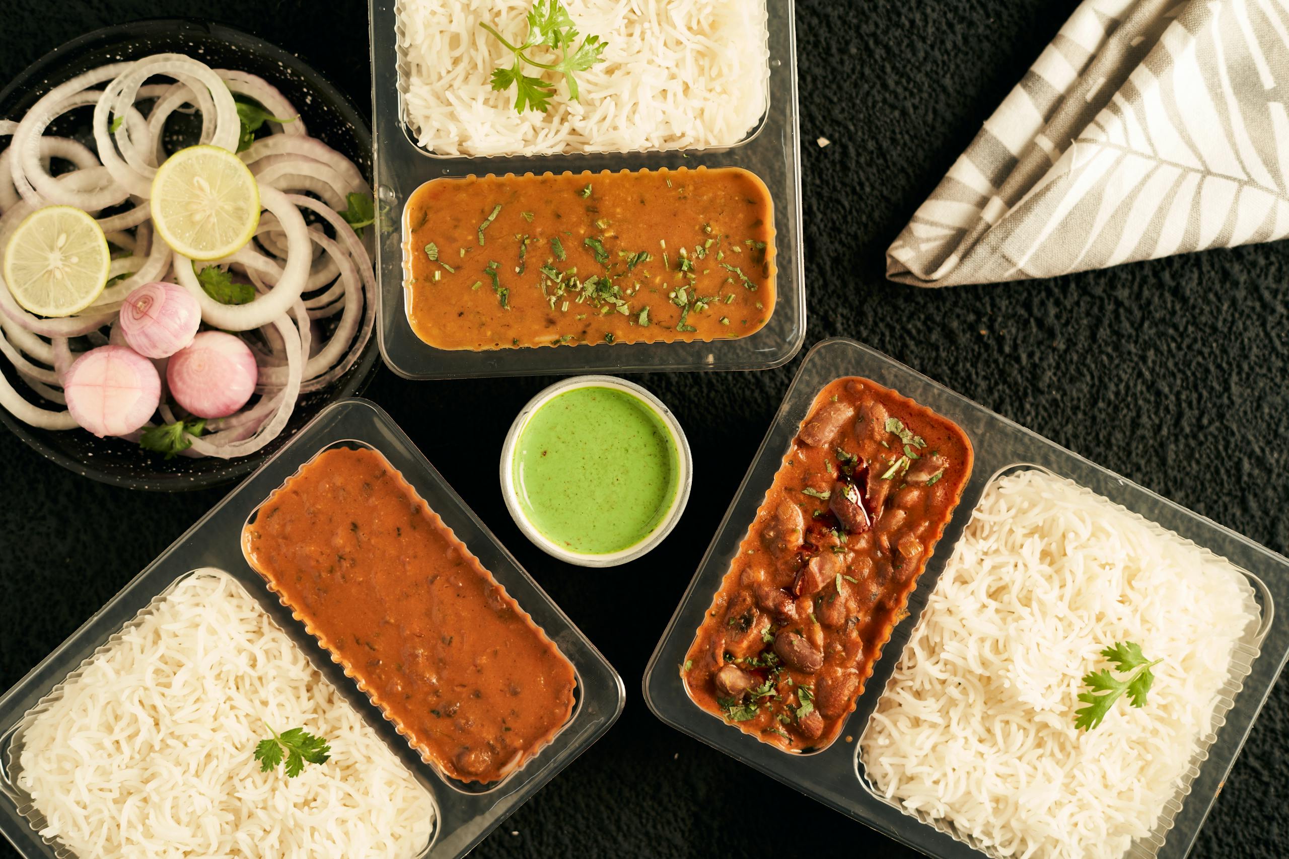 Top view of Indian curry, rice, and greens in takeaway containers with sliced onions.