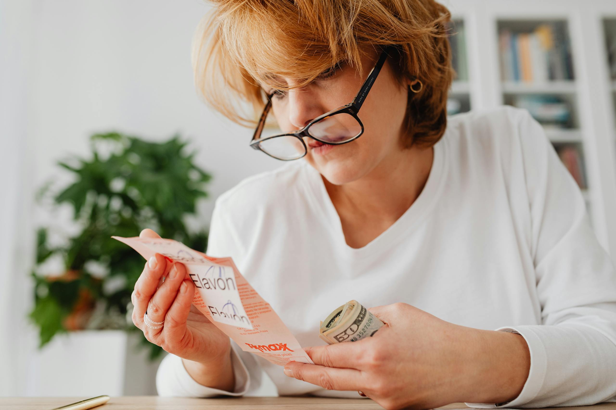 Woman with glasses examines bills and rolls of cash at a desk indoors.