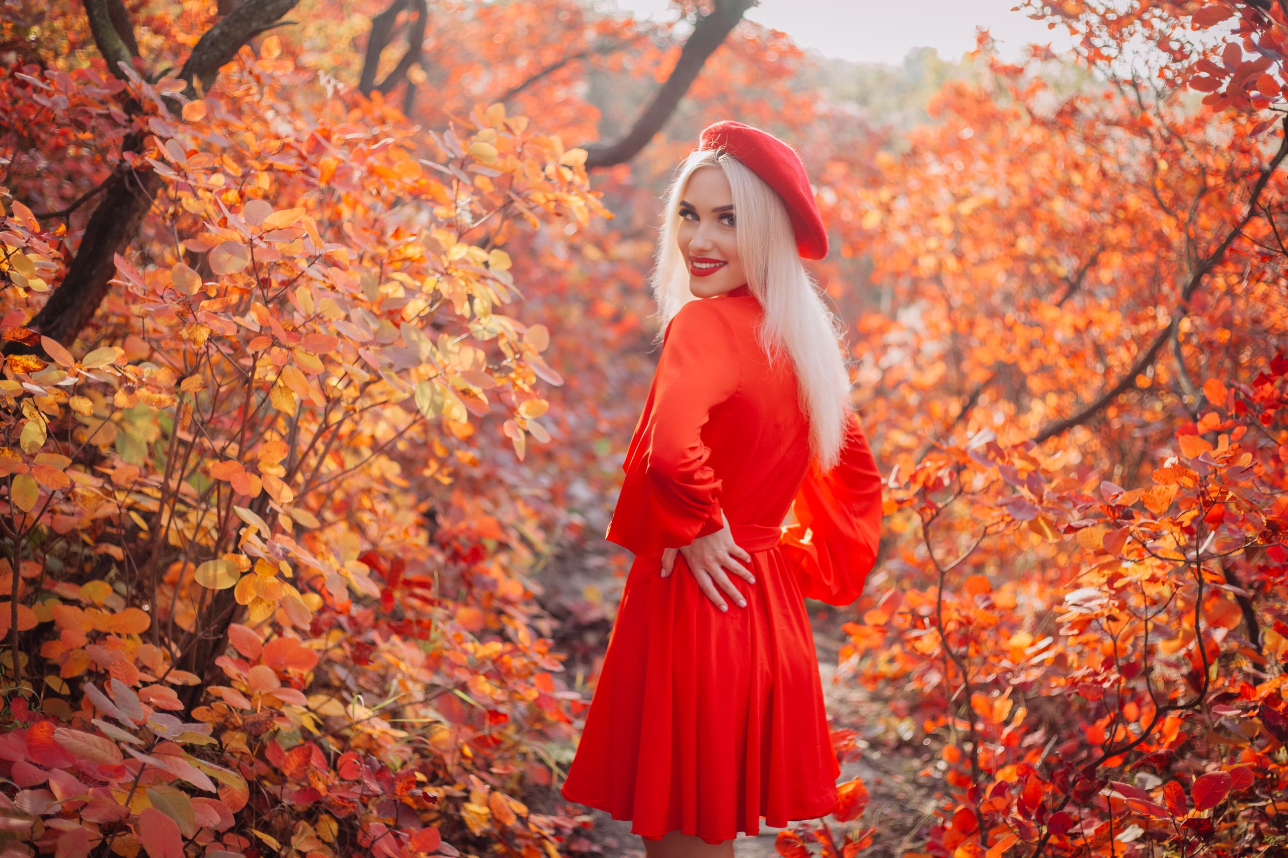 A woman in a red dress poses in a forest with vivid autumn leaves.