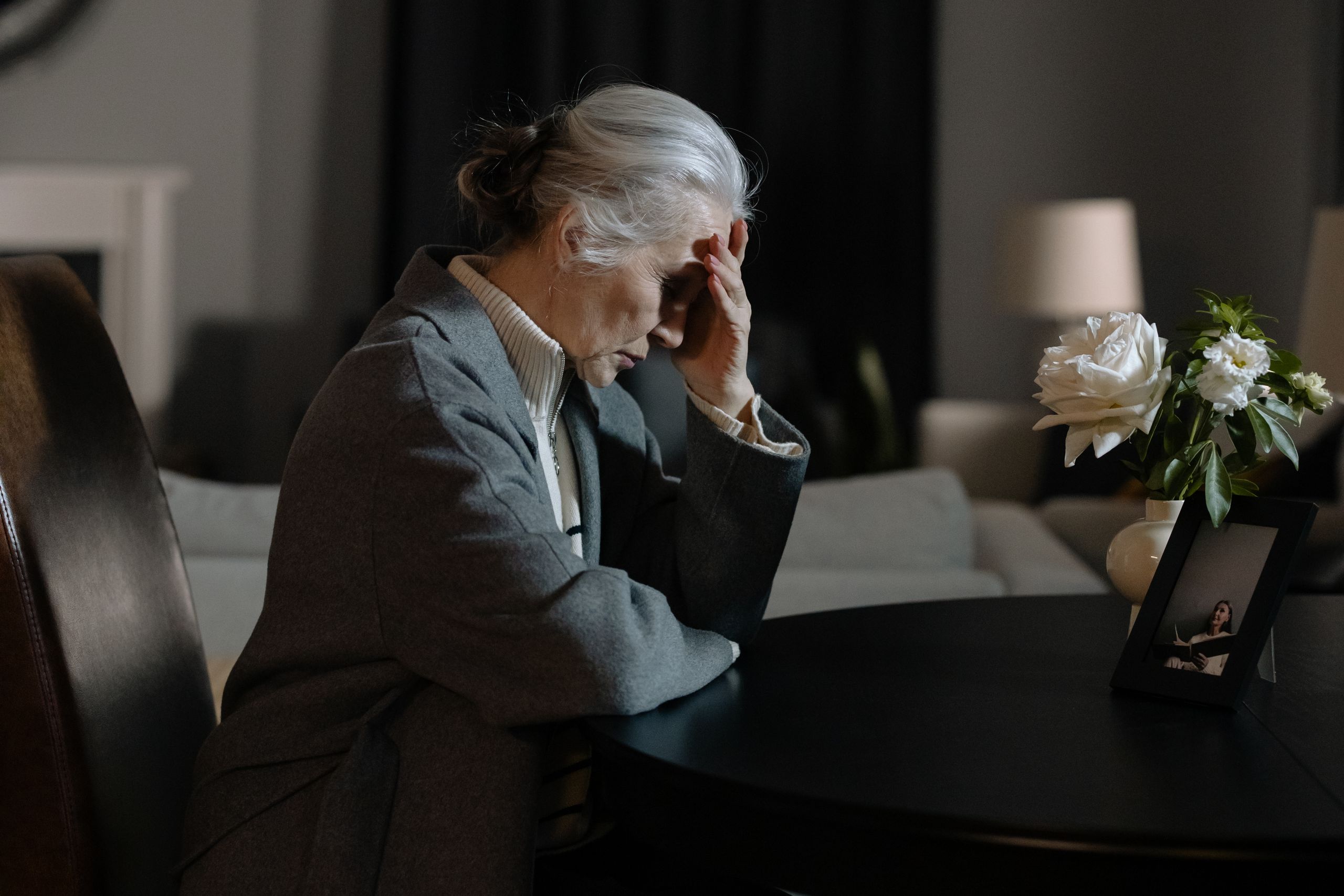 An elderly woman sitting indoors, hand on face, next to a bouquet and framed photo, evokes a reflective mood.