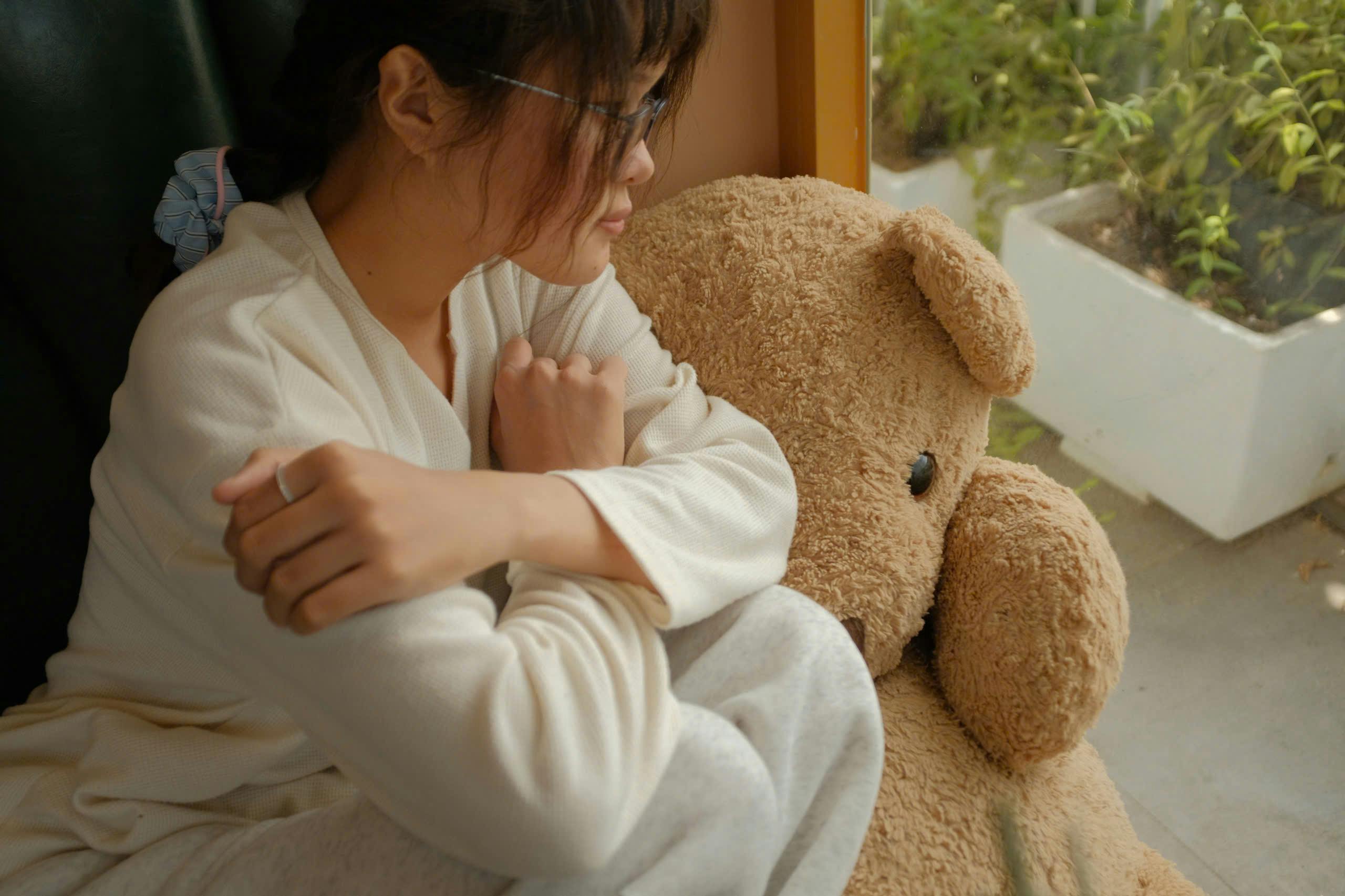 Asian woman in white pajamas, cuddling a teddy bear by a window, indoors.