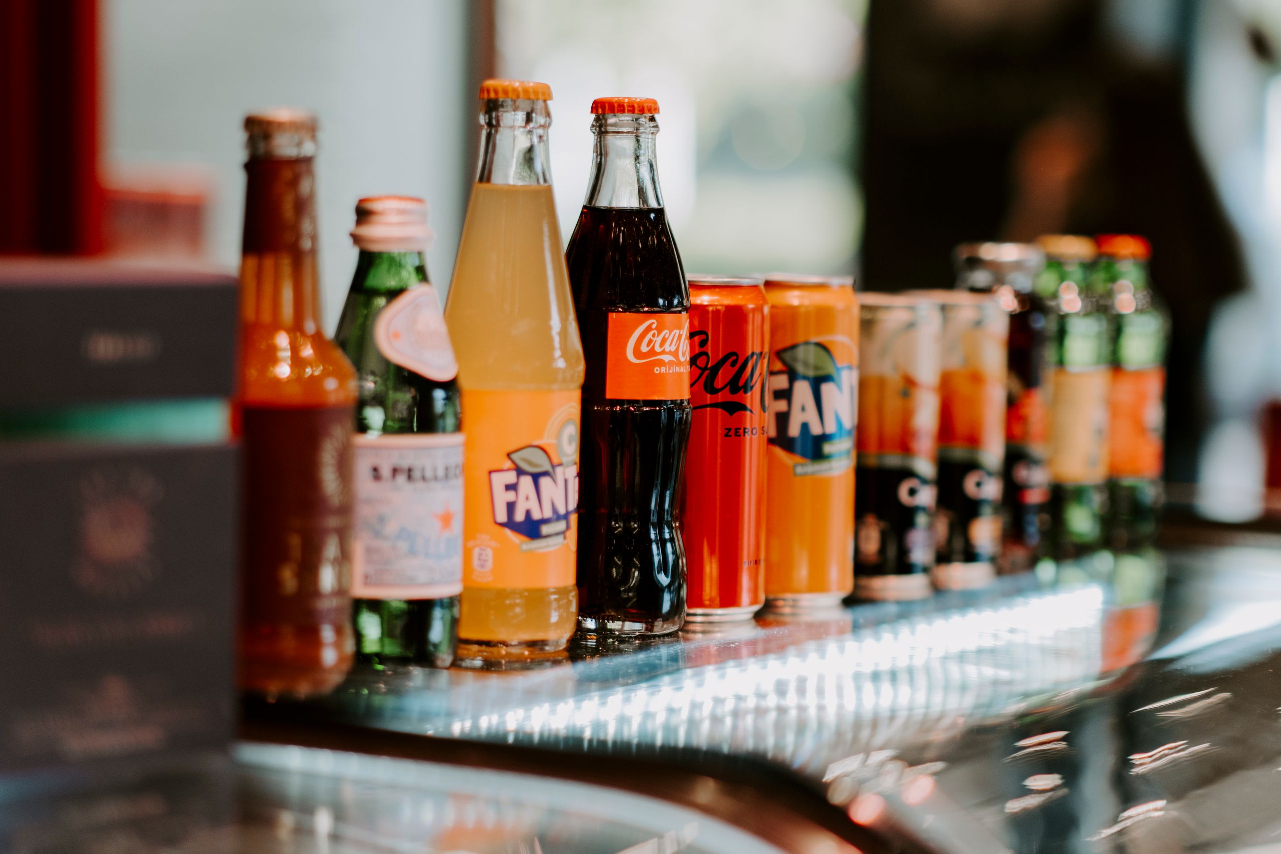 Close-up of assorted bottles and cans of soft drinks, including Coca-Cola and Fanta, on a reflective surface.