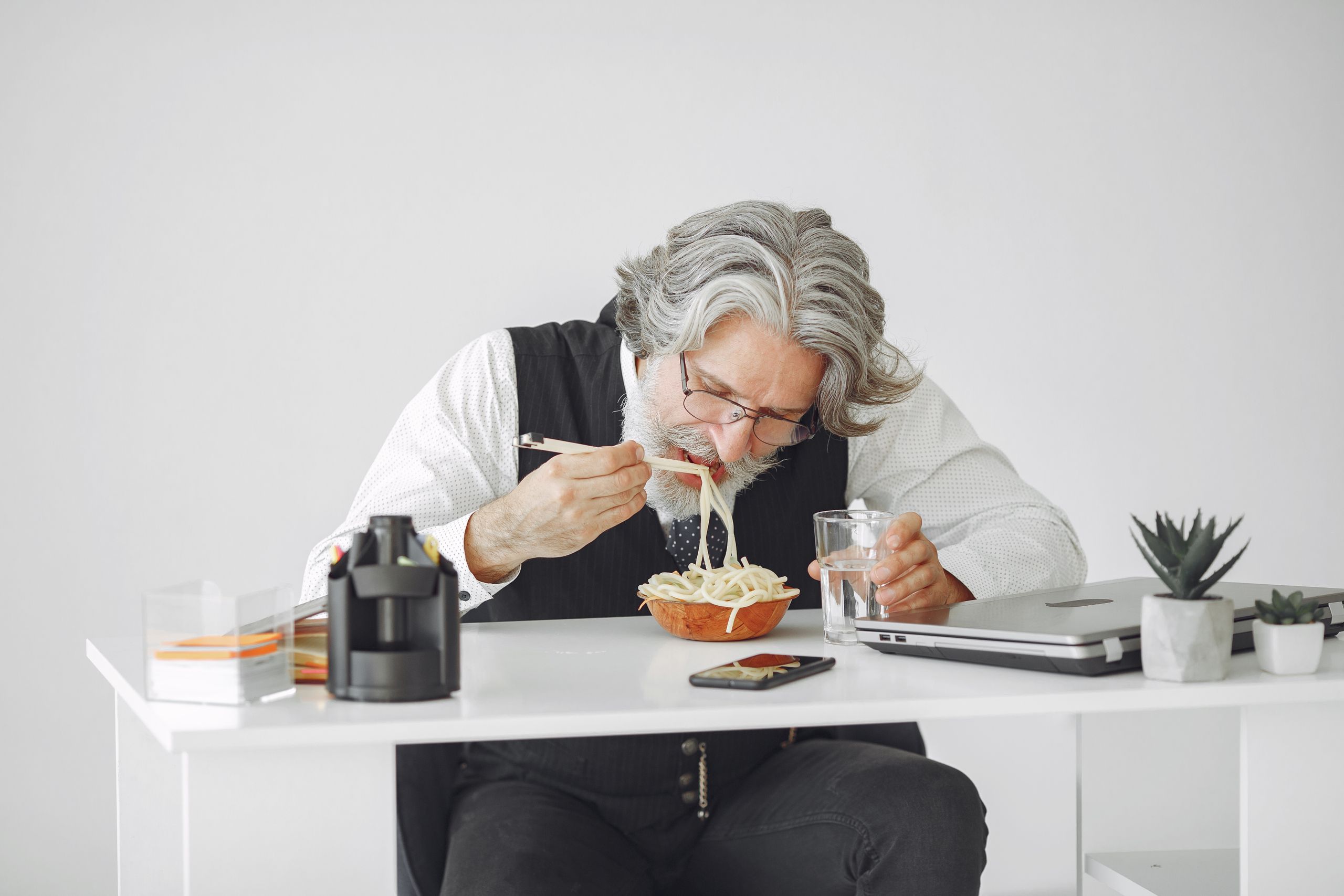 Elderly man enjoying a bowl of noodles while working at a desk with a laptop and plants.