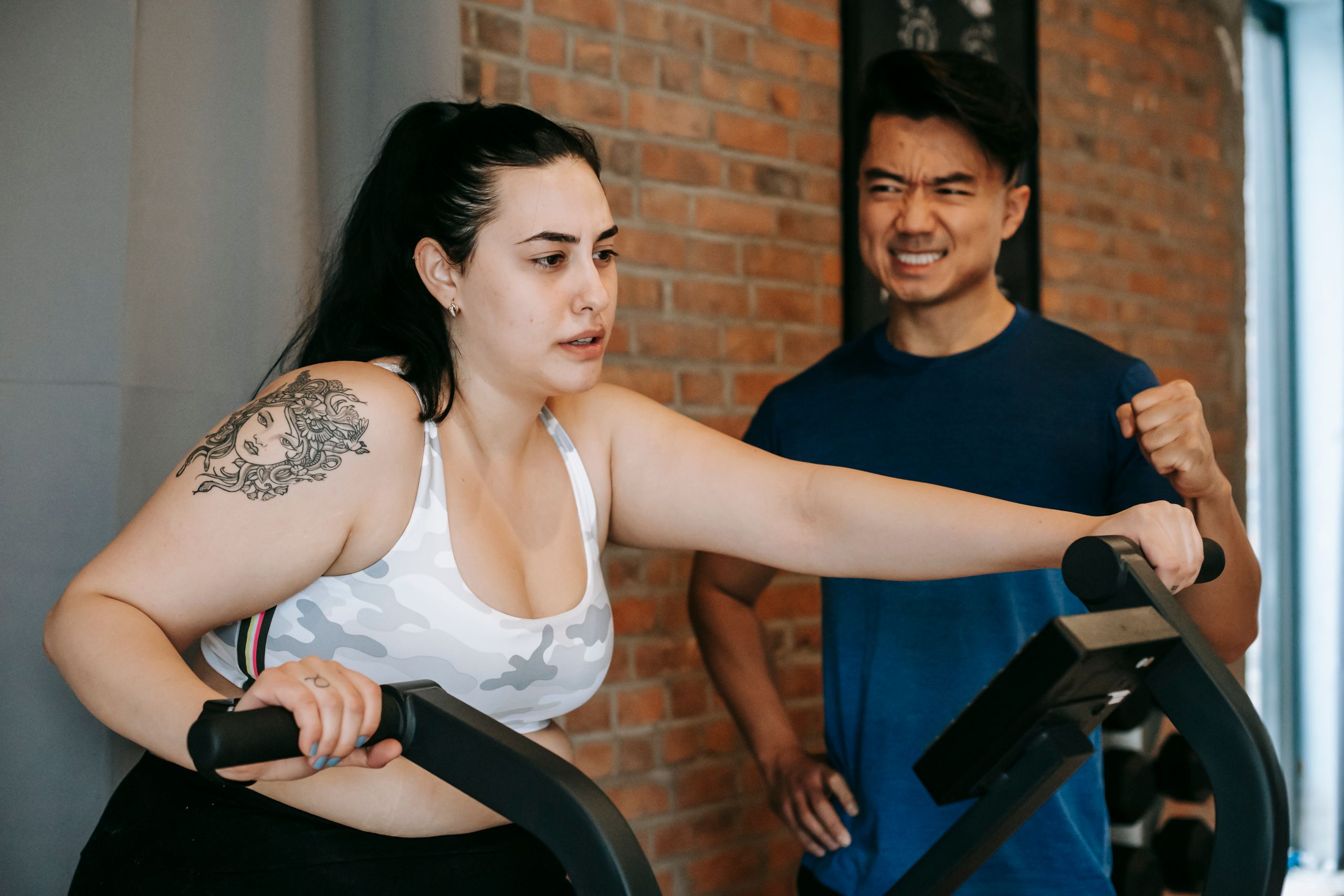 Positive young ethnic male trainer motivating tired tattooed plus size female elliptical machine in modern sports club