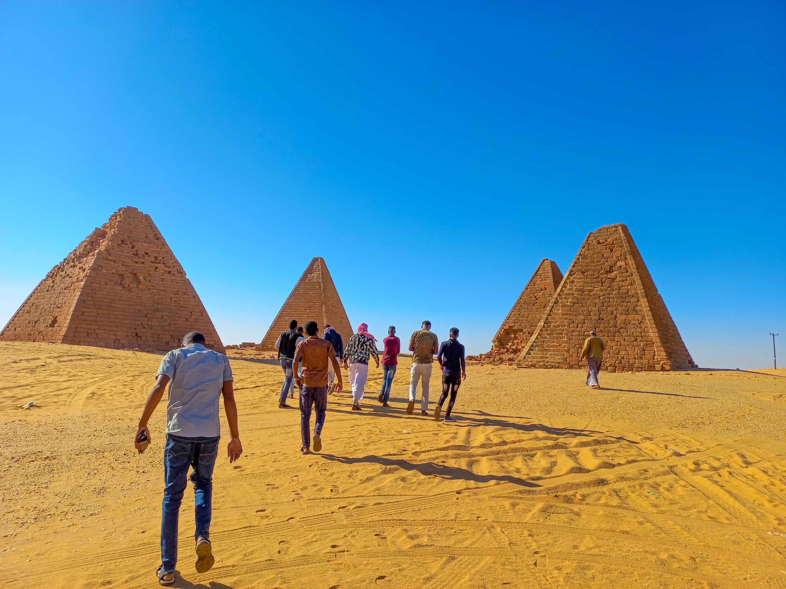 Tourists walk towards ancient pyramids in the Sudanese desert under a clear blue sky.