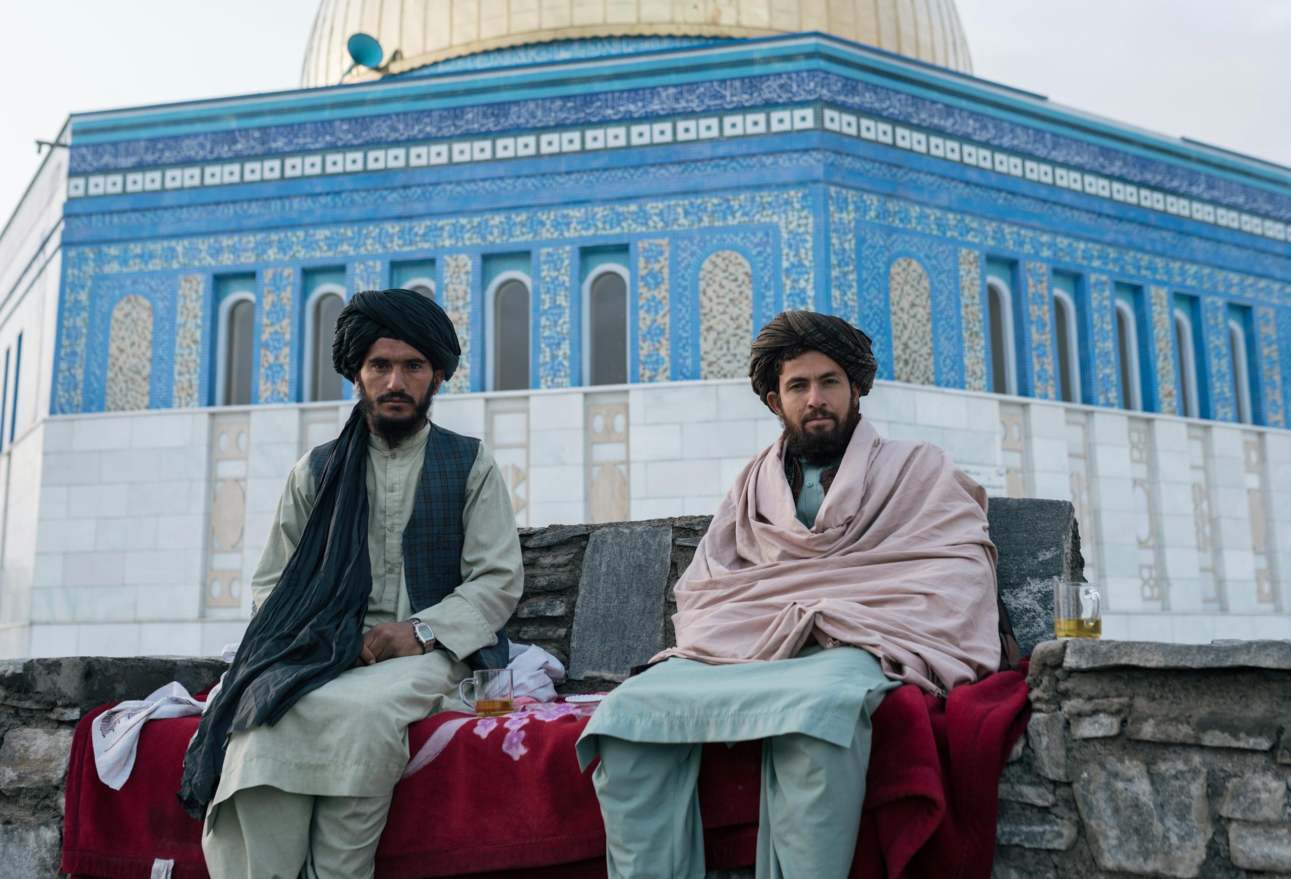 Two Afghan men sit in front of a blue-tiled building, showcasing traditional attire in Afghanistan.