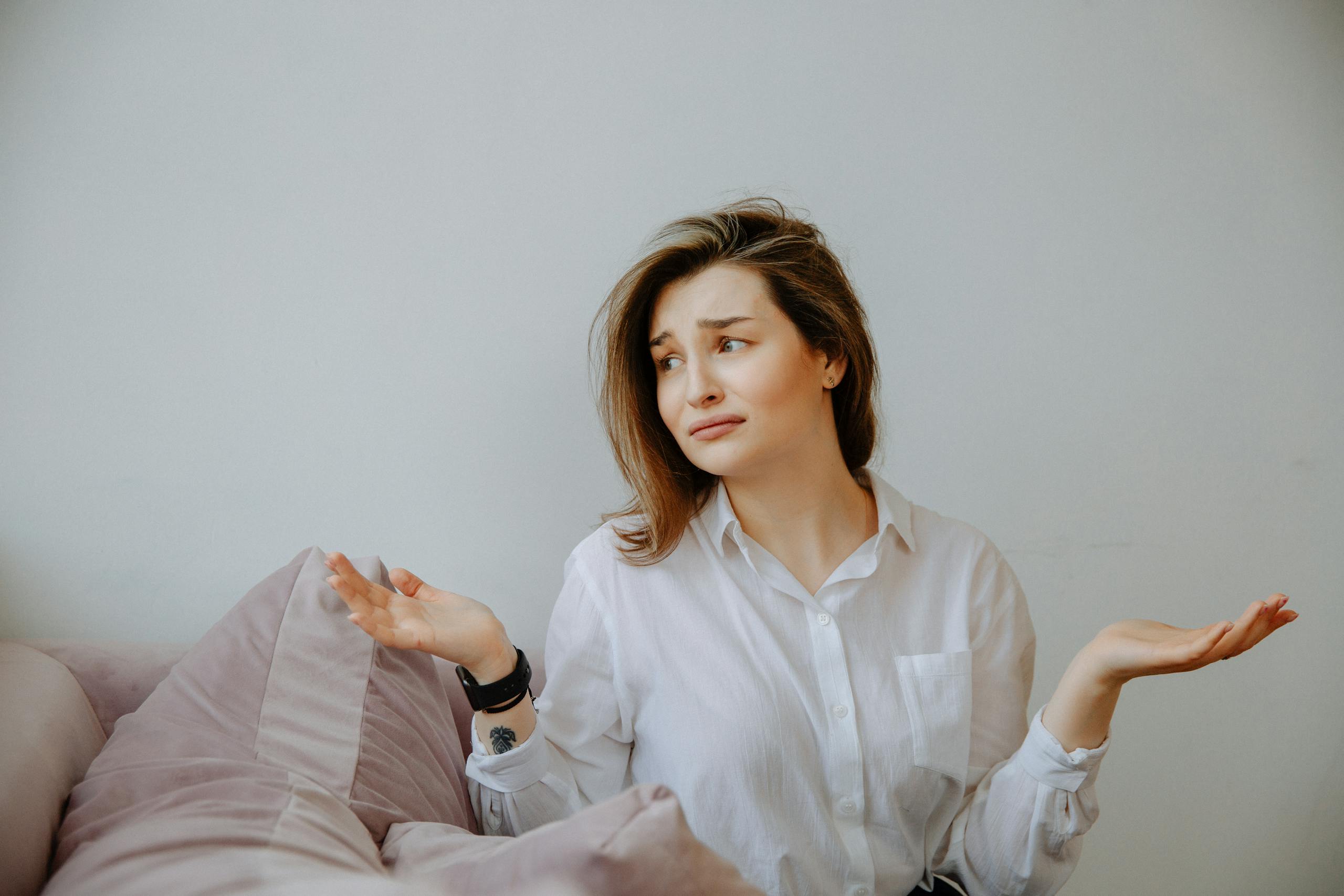 Woman in white shirt sitting indoors on a couch, gesturing with a confused expression, hands up in a shrug.