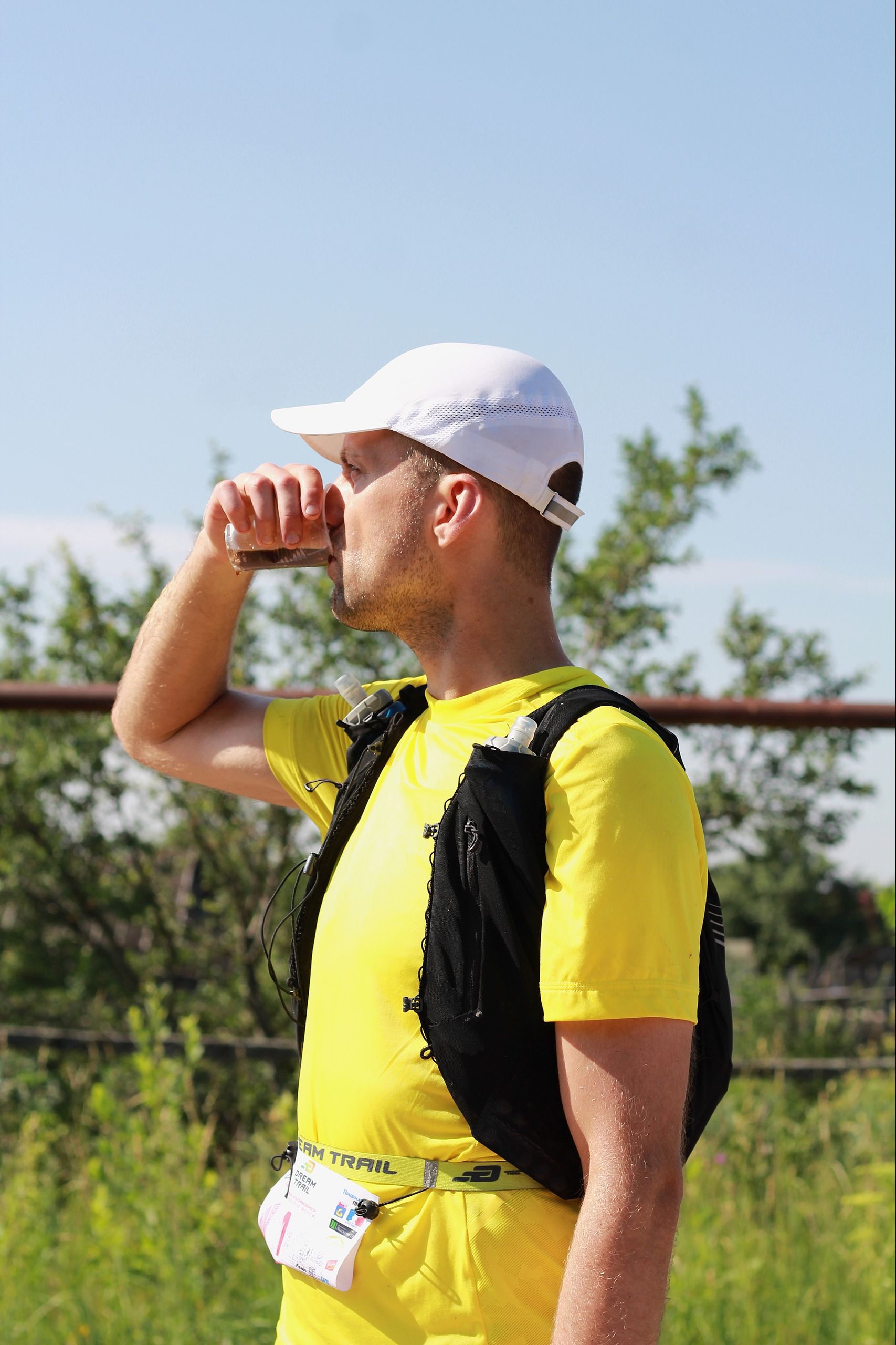 Man in yellow shirt drinks water from bottle during outdoor run on a sunny day.