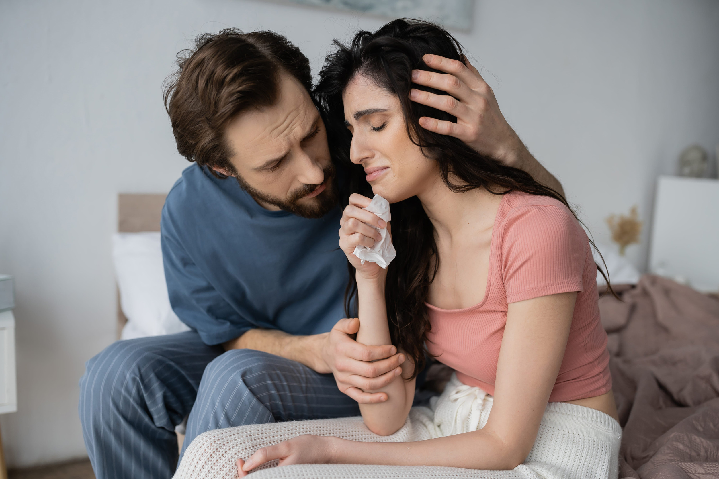 Caring man in pajama hugging crying and upset girlfriend on bed at home
