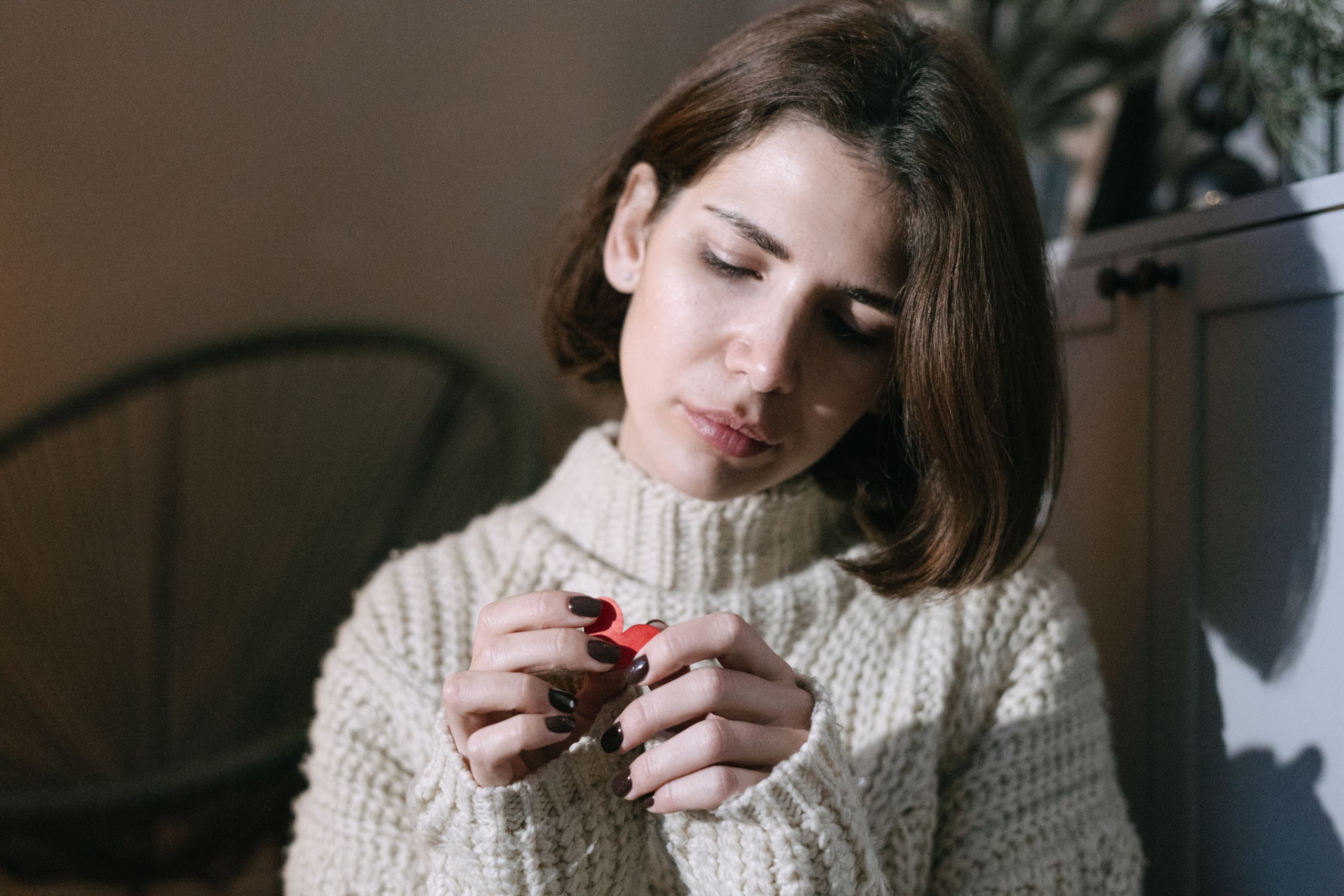 A brunette woman holding a red paper heart, looking thoughtful in a cozy indoor setting.