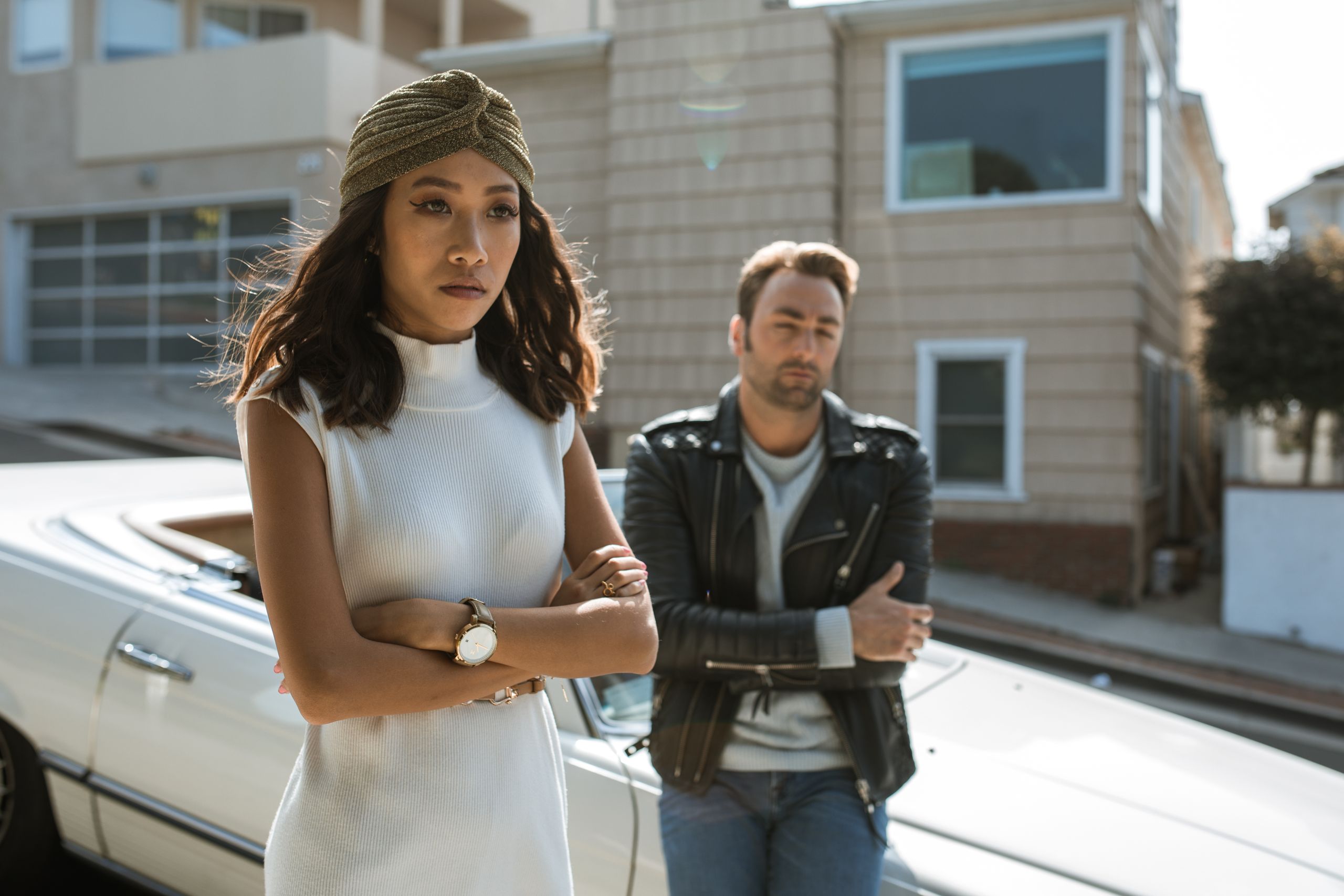 A couple appears upset, standing apart on a sunny urban street near a vintage car.