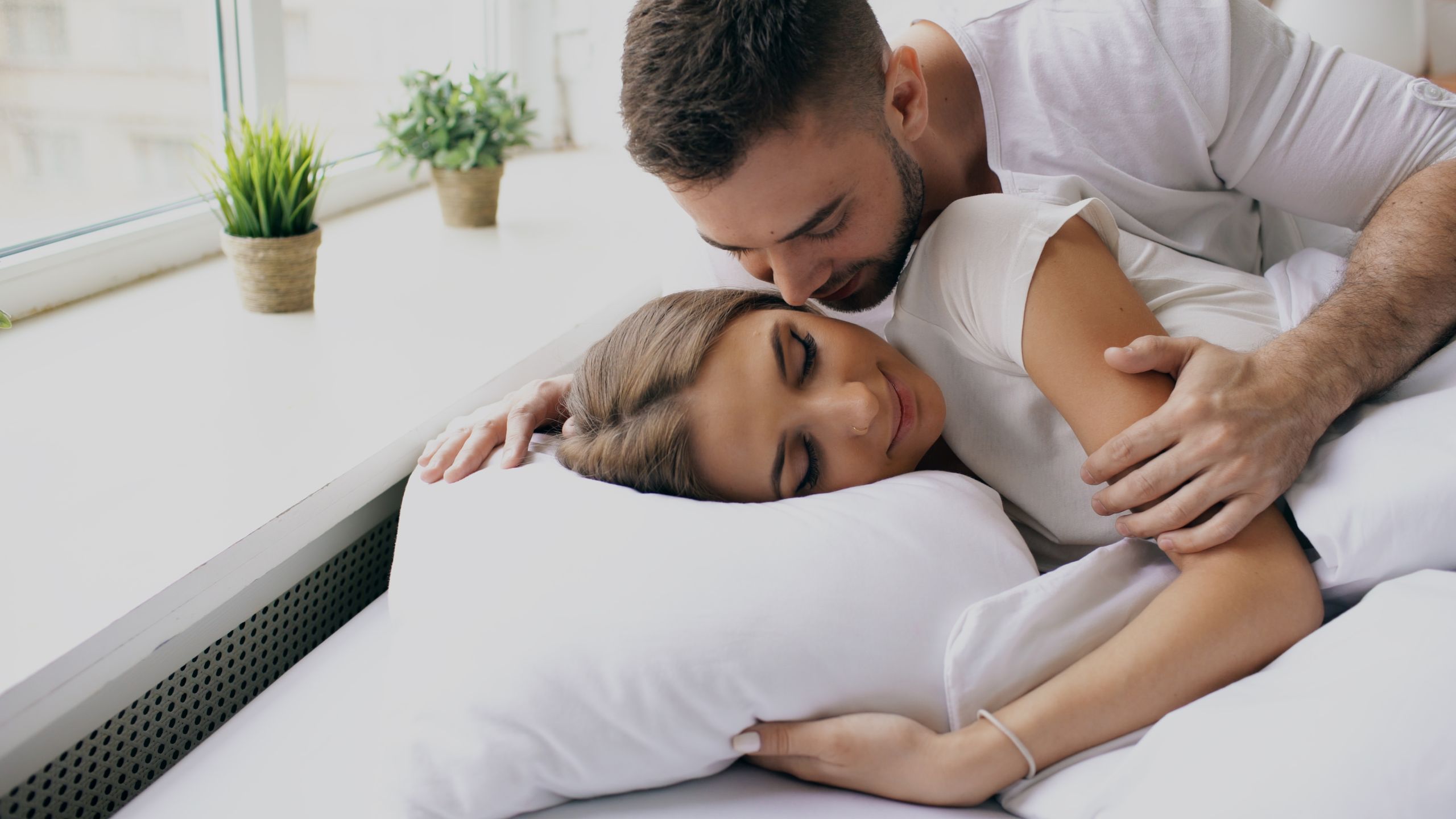 A loving couple cuddling in bed by a large window, embracing intimacy.