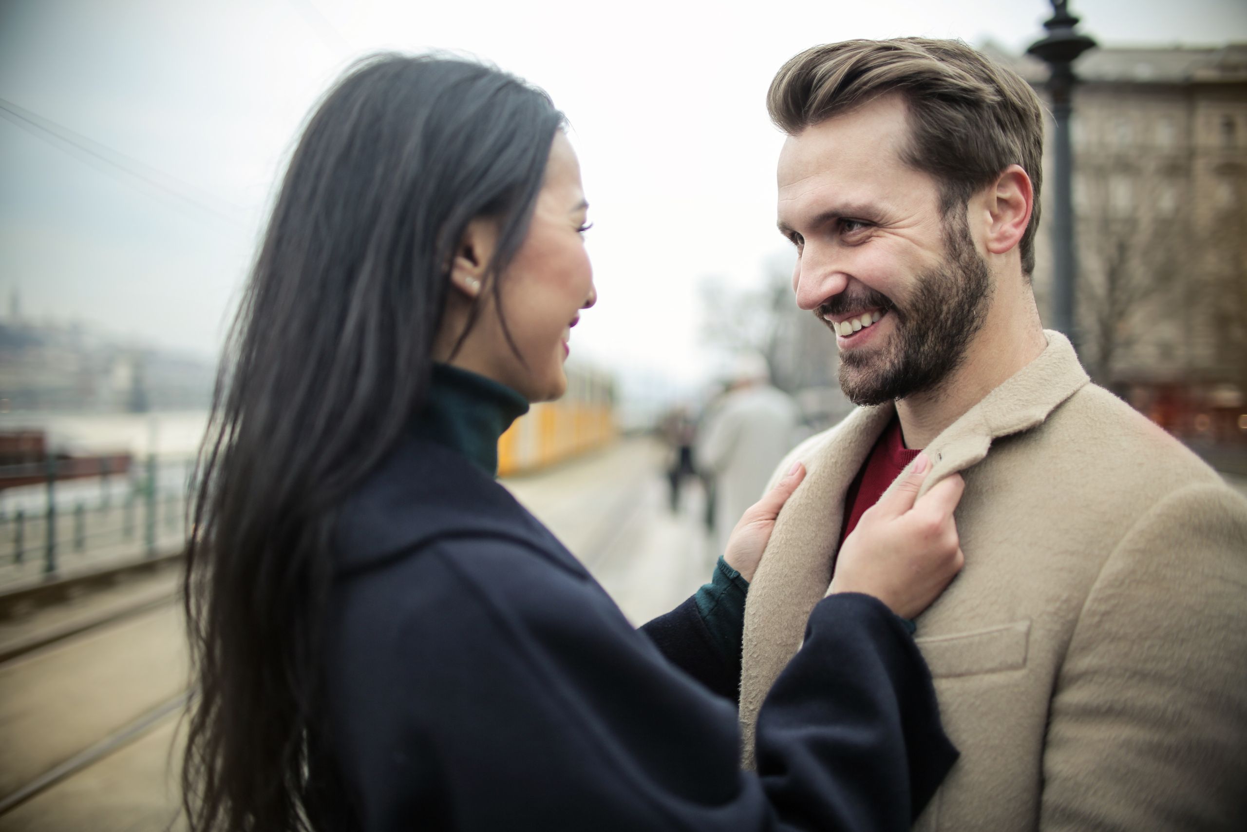 A smiling couple in a city street, sharing a moment of joy and affection outdoors on a cloudy day.