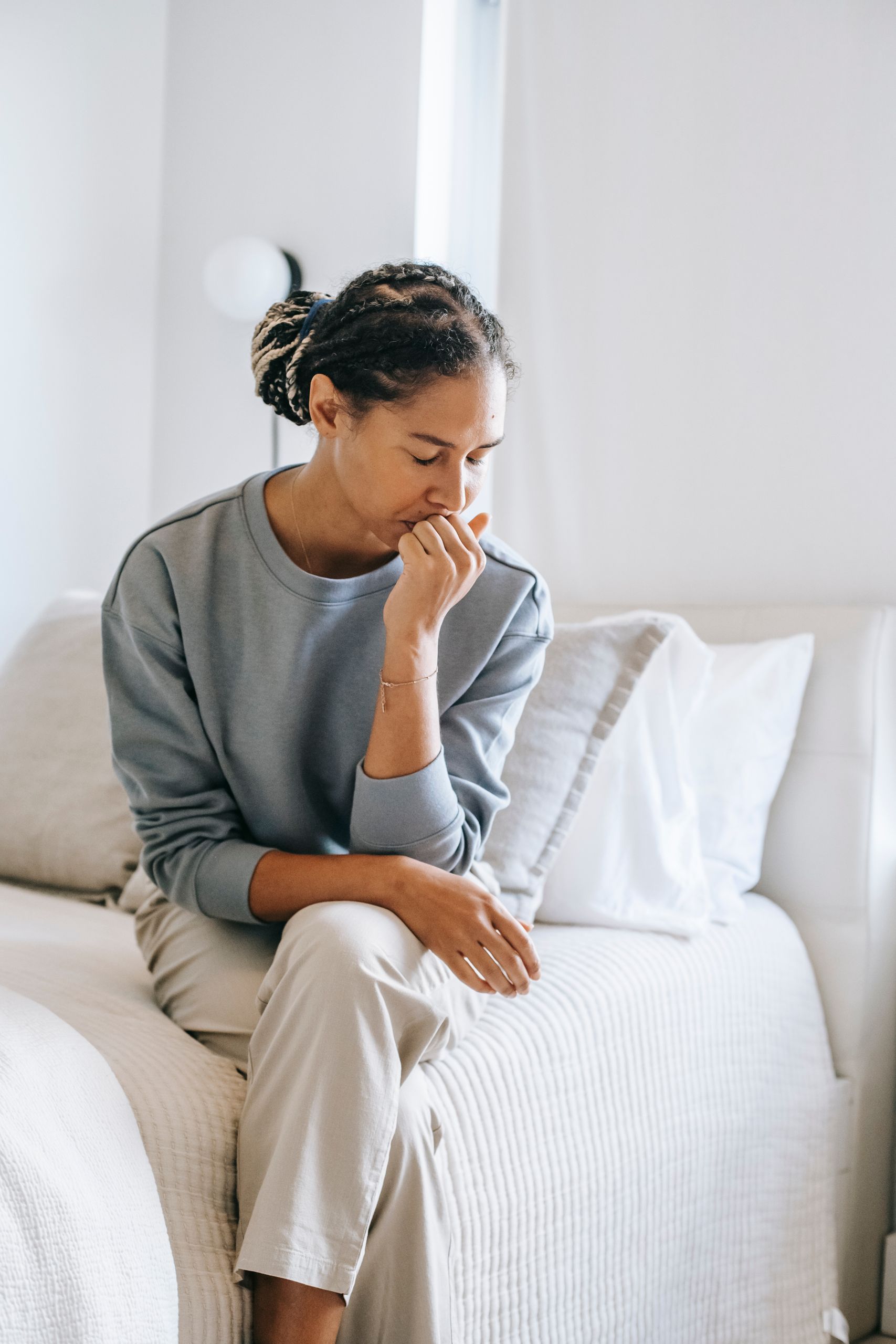 Sorrowful crying African American female sitting with eyes closed and crossed legs on bed and touching lips in stress