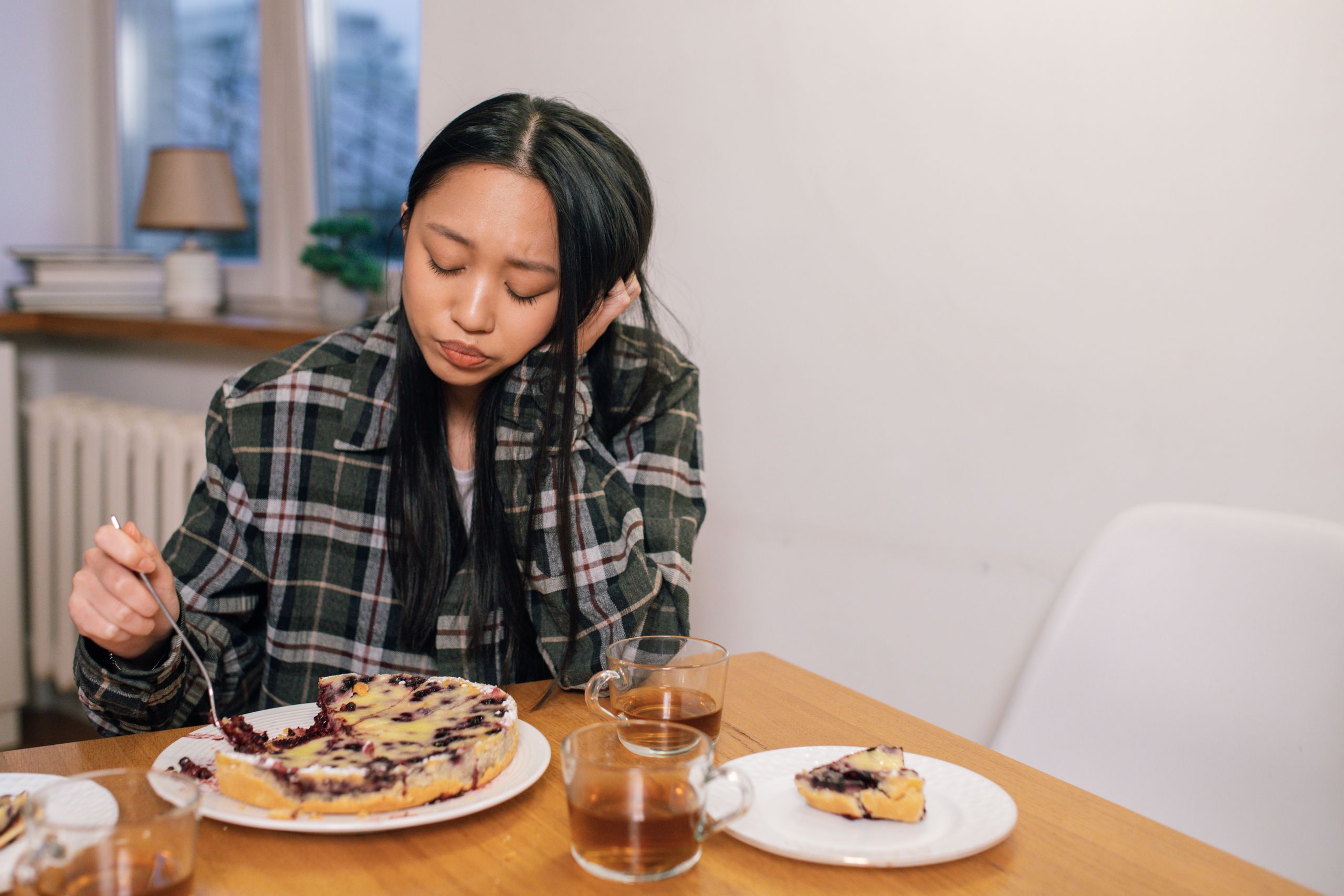 Woman in plaid shirt thoughtfully eats blueberry pie in a cozy indoor setting.