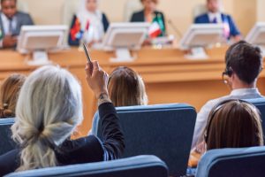 Back view of audience in armchairs raising their hands to ask questions to speaker after report at conference