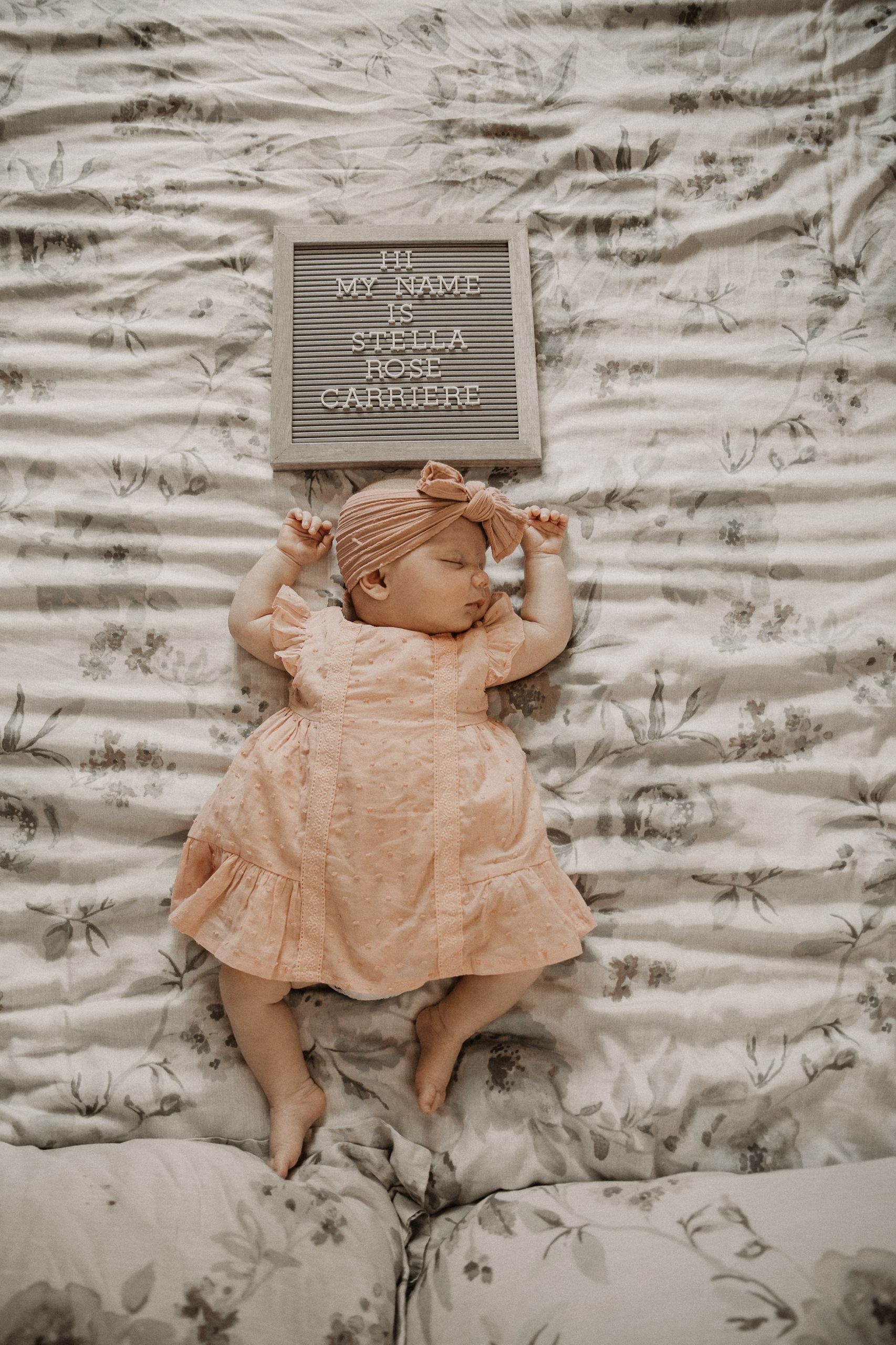 Adorable newborn baby girl sleeping peacefully with a name board nearby, captured in a soft and pastel setting.