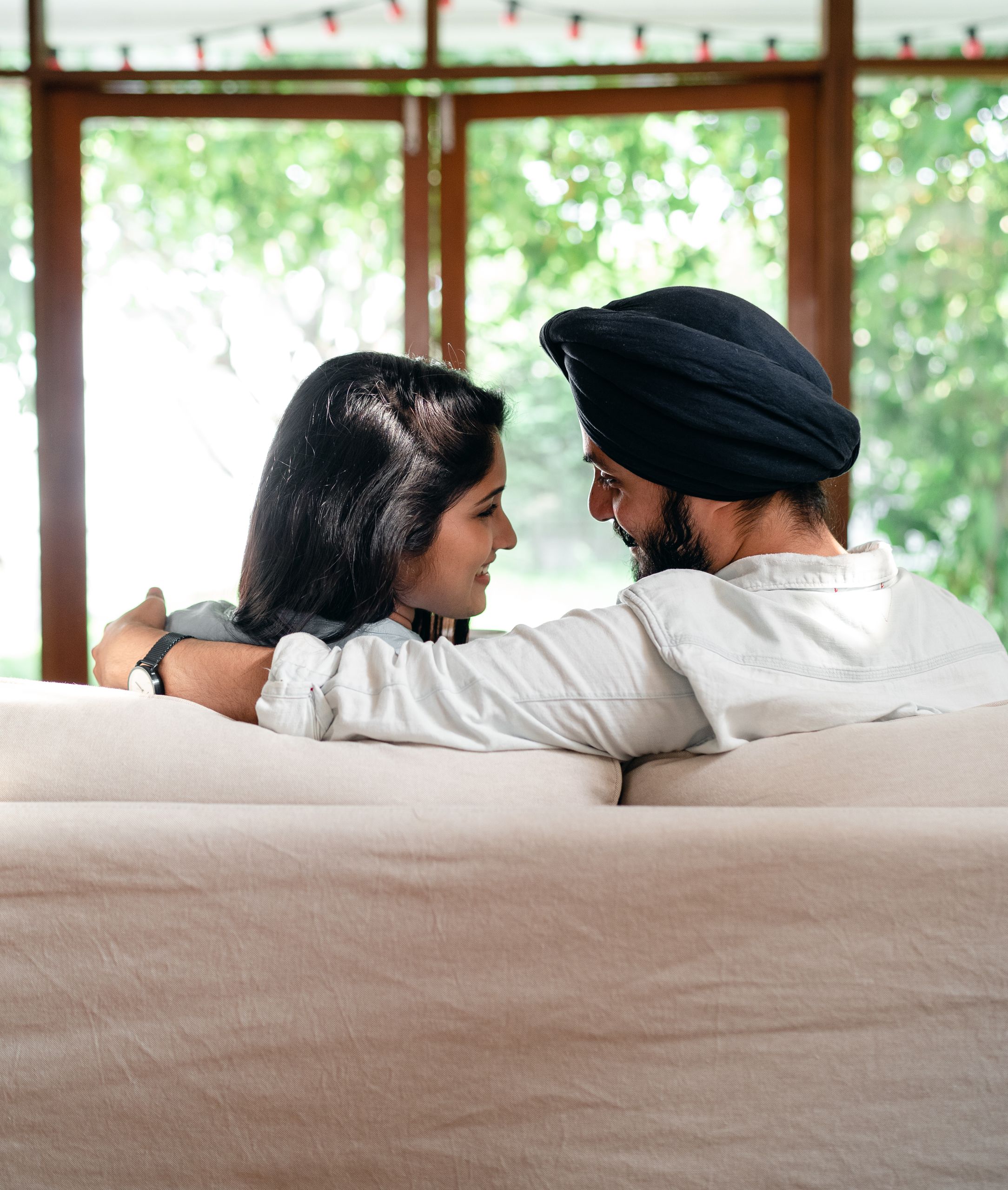 Back view of happy romantic young Indian couple cuddling while relaxing on coach in cozy apartment and looking at each other