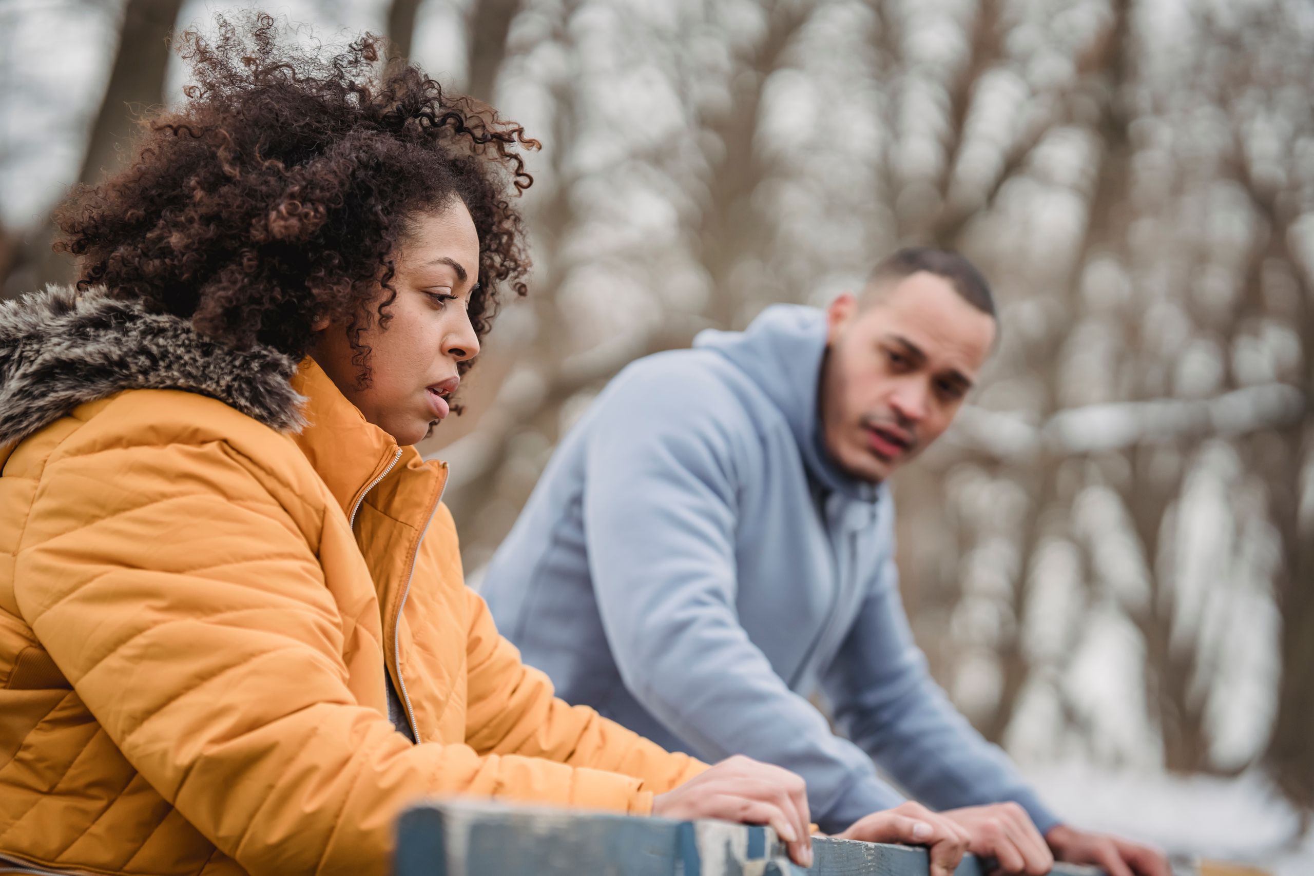 Determined African American man and woman in warm clothes leaning on wooden barrier during workout in winter park on blurred background