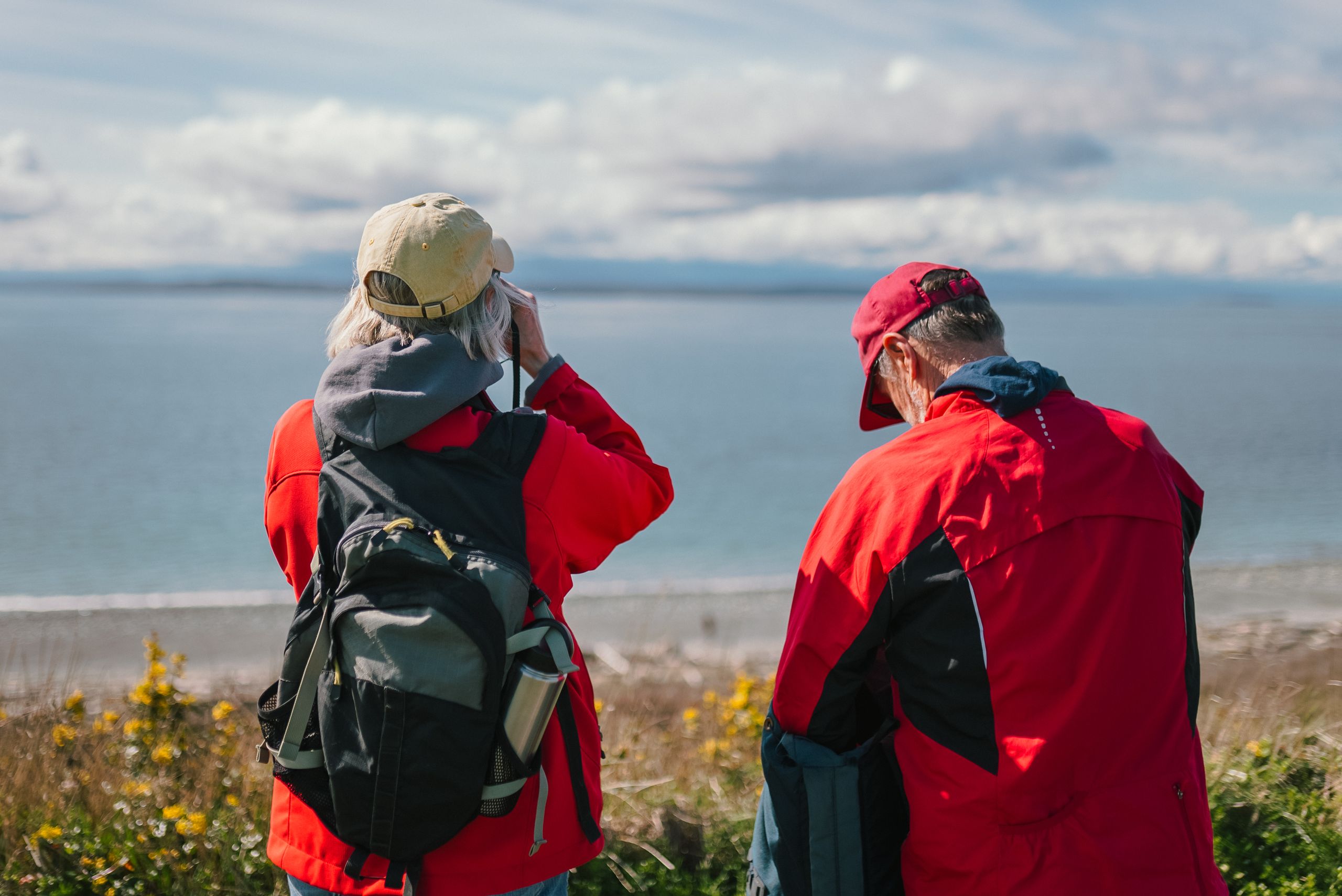 Elderly couple in red jackets enjoying seaside view, outdoors with backpacks.