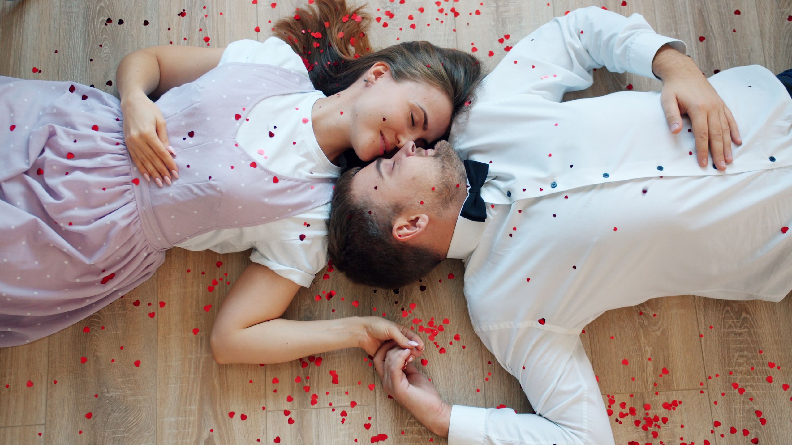 Happy couple lying on the floor surrounded by heart confetti, embracing love.