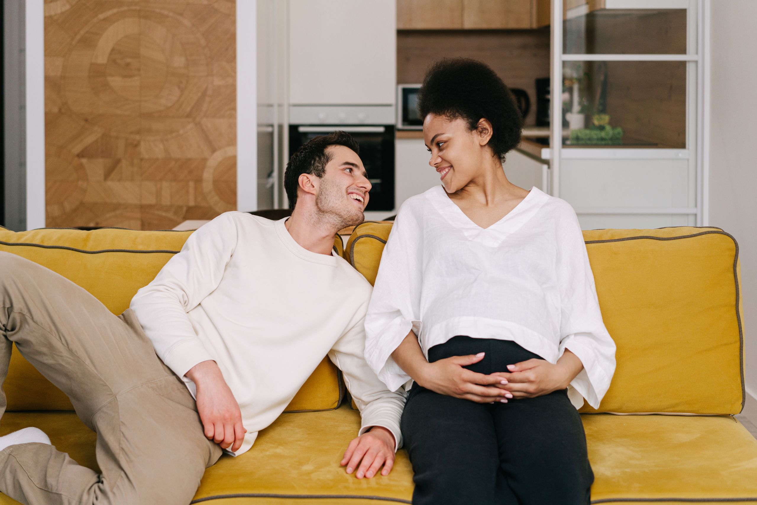 Joyful expectant couple relaxing at home on a vibrant yellow couch, sharing a loving moment.