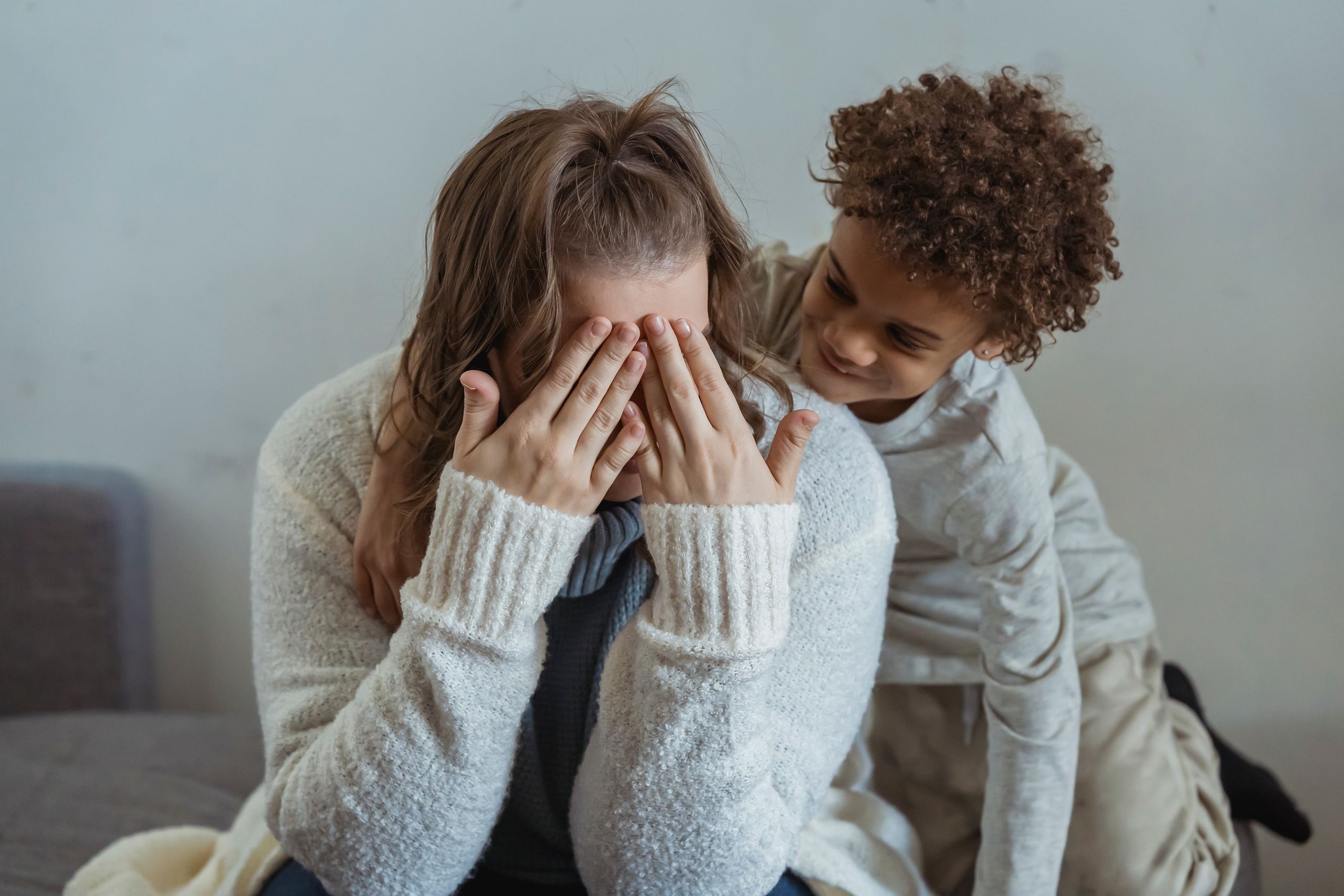 Mother covering face with hands while playing with ethnic kid on sofa in light room