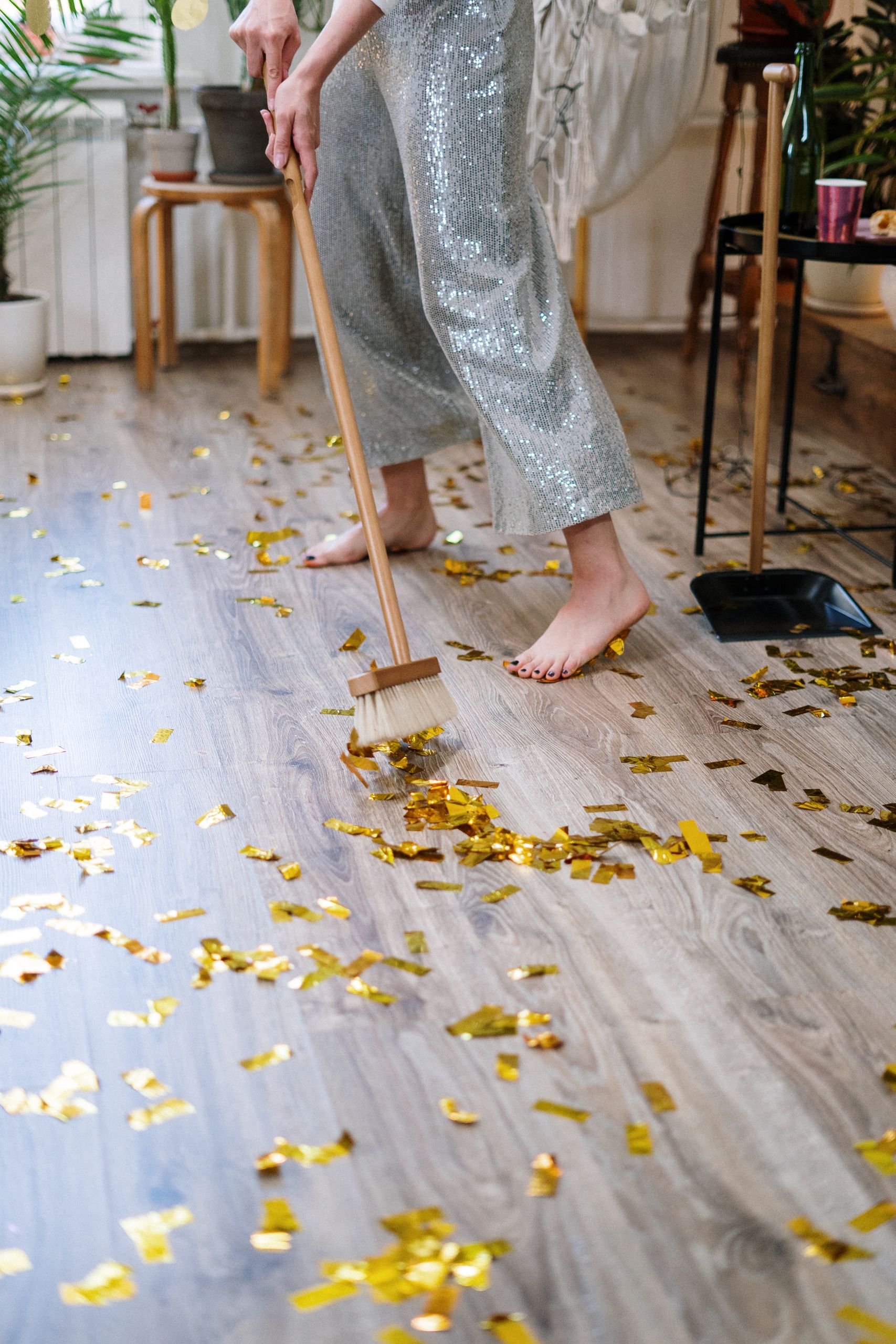 Person sweeping golden confetti off wooden floor after a party, barefoot indoors.