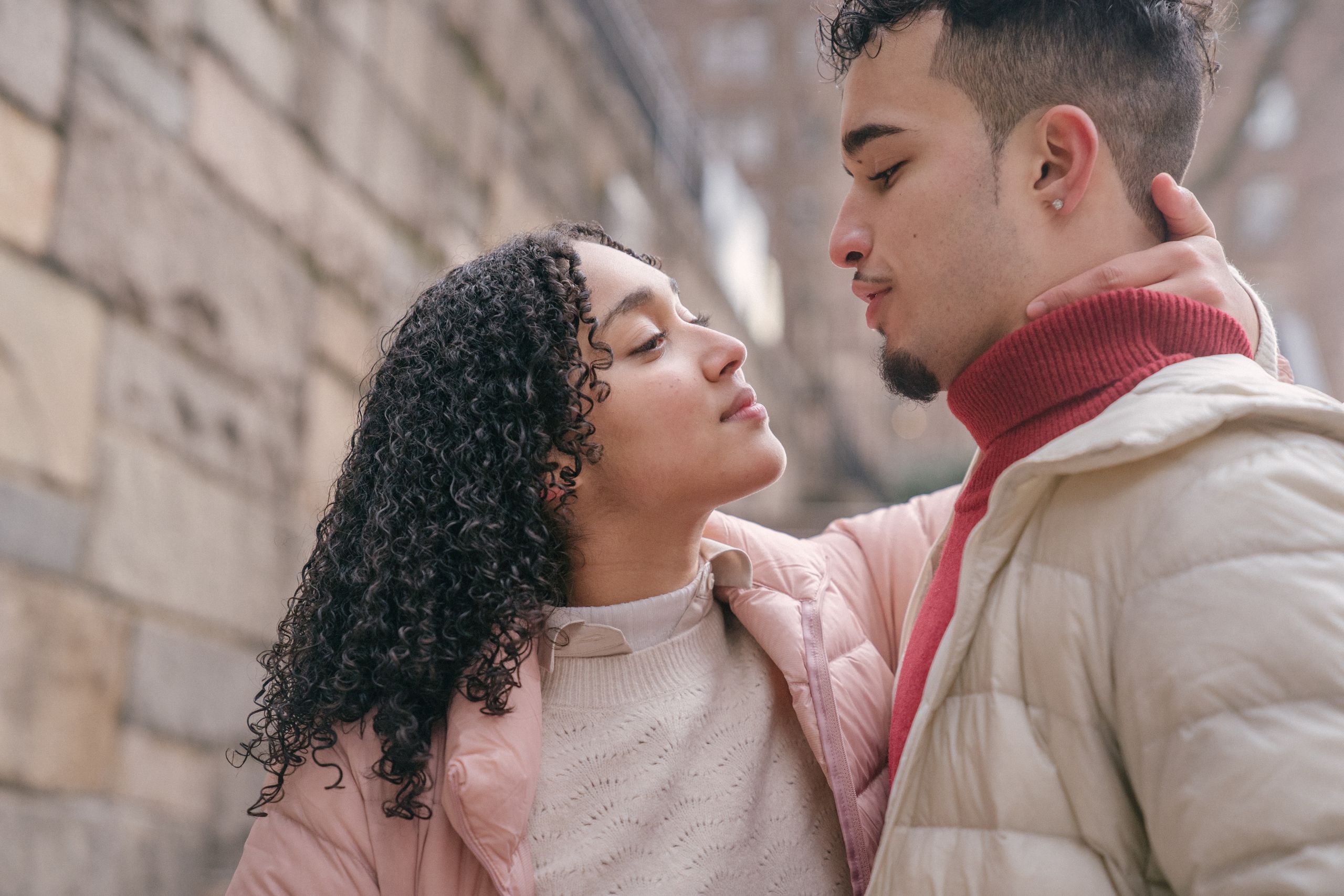 Side view of tender Hispanic couple looking at each other while embracing on street near stone wall on street on blurred background
