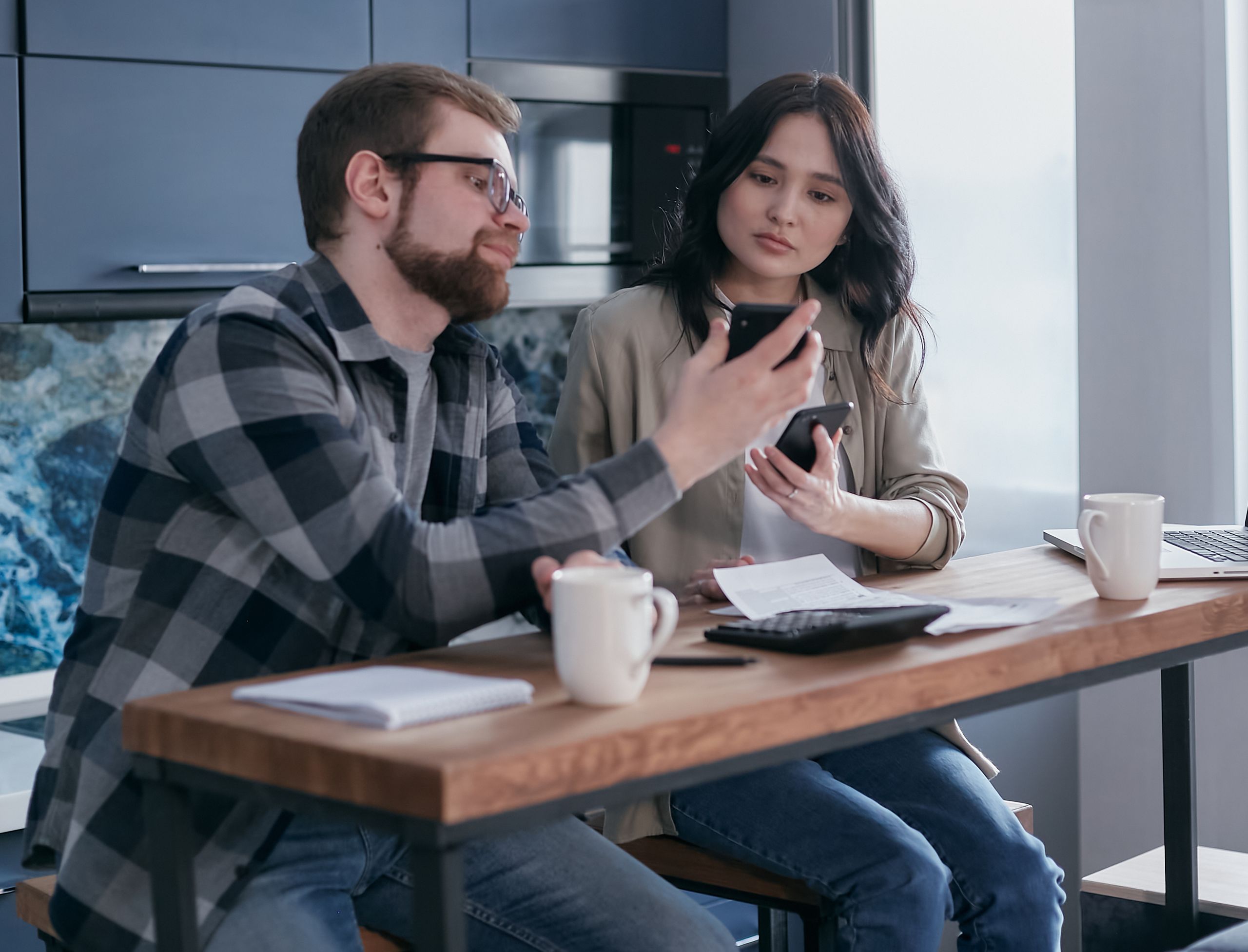 Young couple discussing finances using mobile phones at a kitchen table.