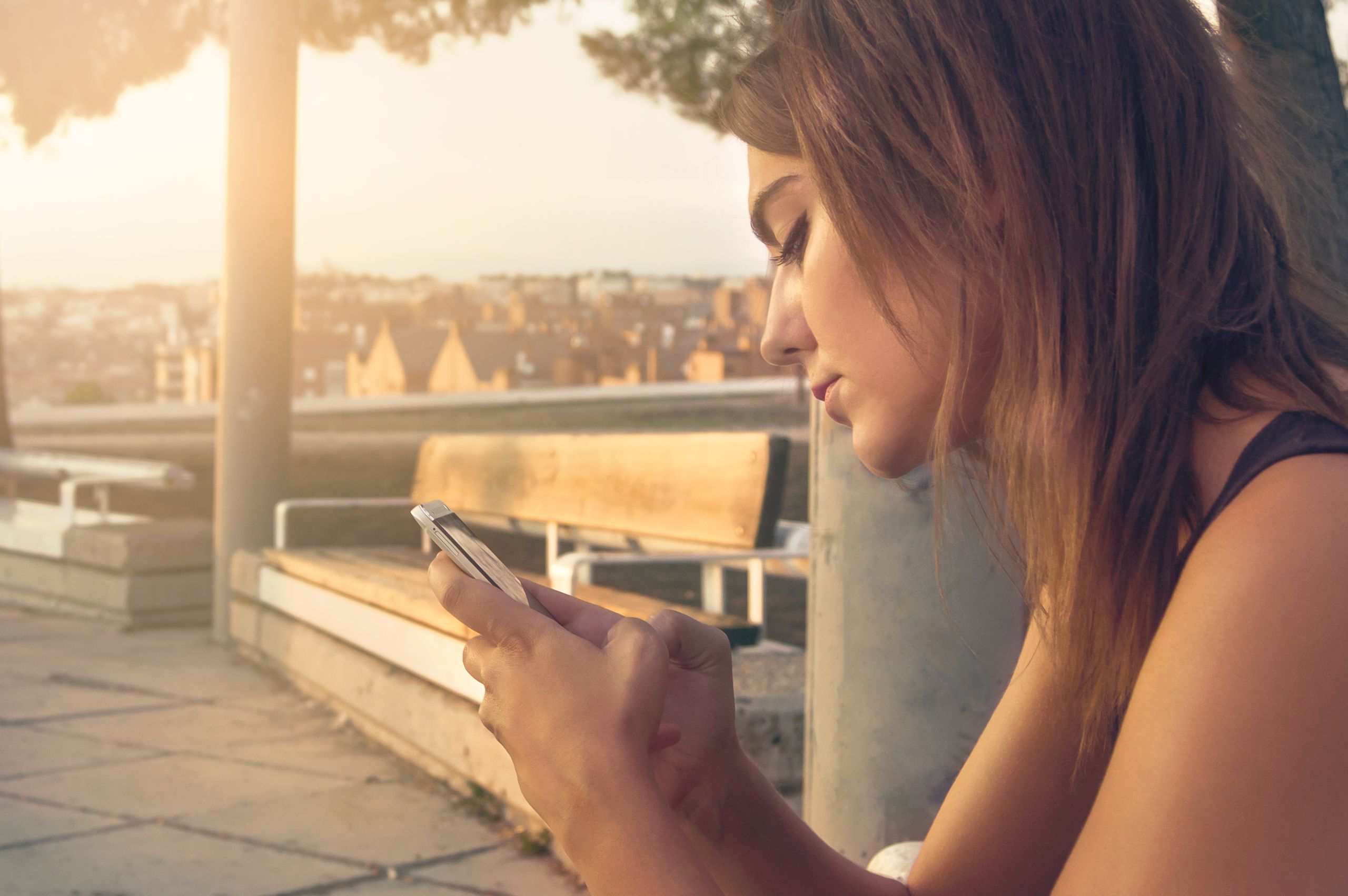 Young woman texting on smartphone while sitting outdoors in sunny park setting.